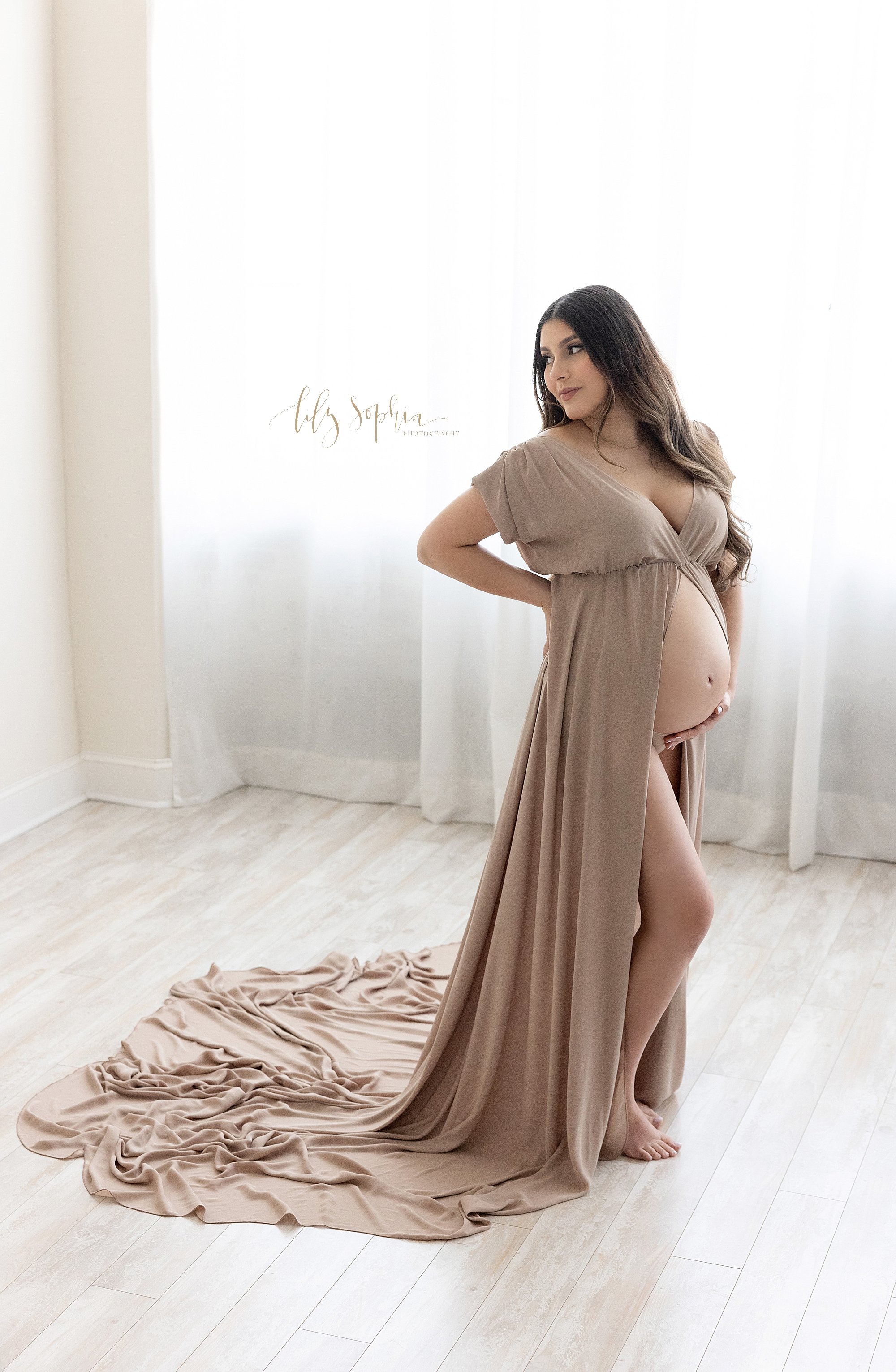  Maternity photo shoot with a pregnant mother wearing a split front short-sleeved V-necked gown as she stands with her left hand on her hip  while looking over her left shoulder and her right hand holding the base of her bare belly taken  in front of