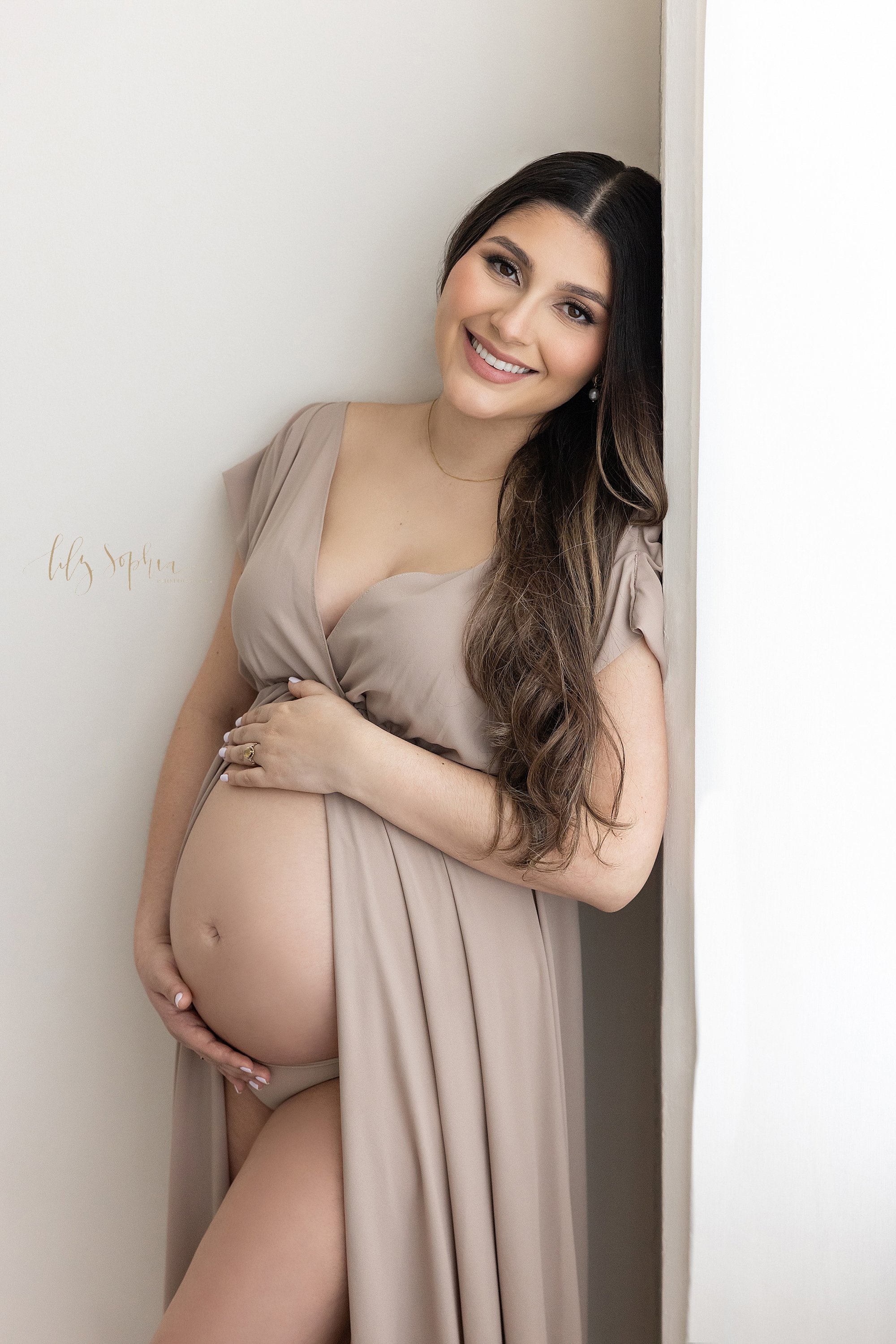  Maternity portrait of a pregnant mother as she leans against a window streaming natural light while she frames her bare belly while wearing a split-front knit gown taken in a photography studio near Morningside in Atlanta. 