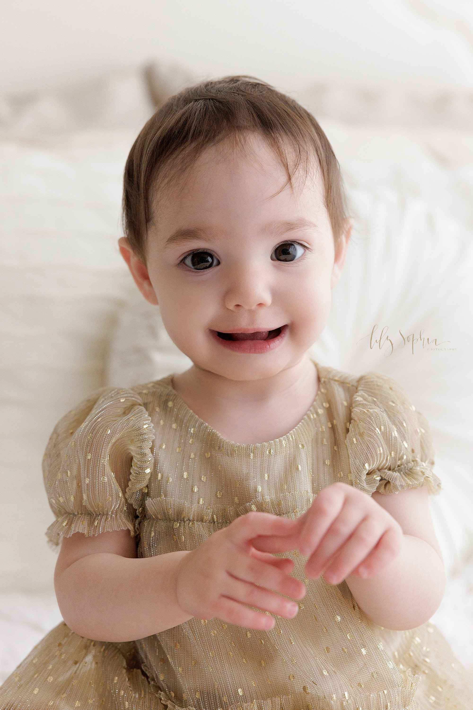  First birthday portrait of a one year old girl as she sits on a bed smiling in her fancy gold dress taken in natural light in a photography studio near Cumming in Atlanta. 