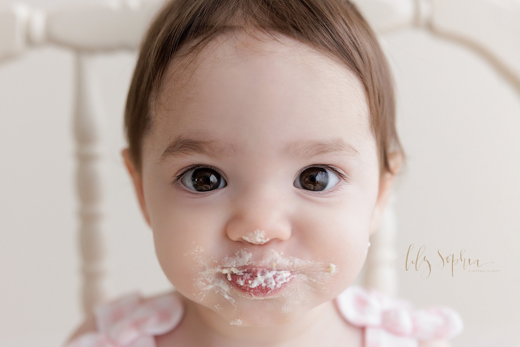  First birthday smash cake photo shoot with a one year old little girl with big brown eyes as she puckers her lips that are covered with icing taken in a natural light photography studio near Roswell in Atlanta, Georgia. 