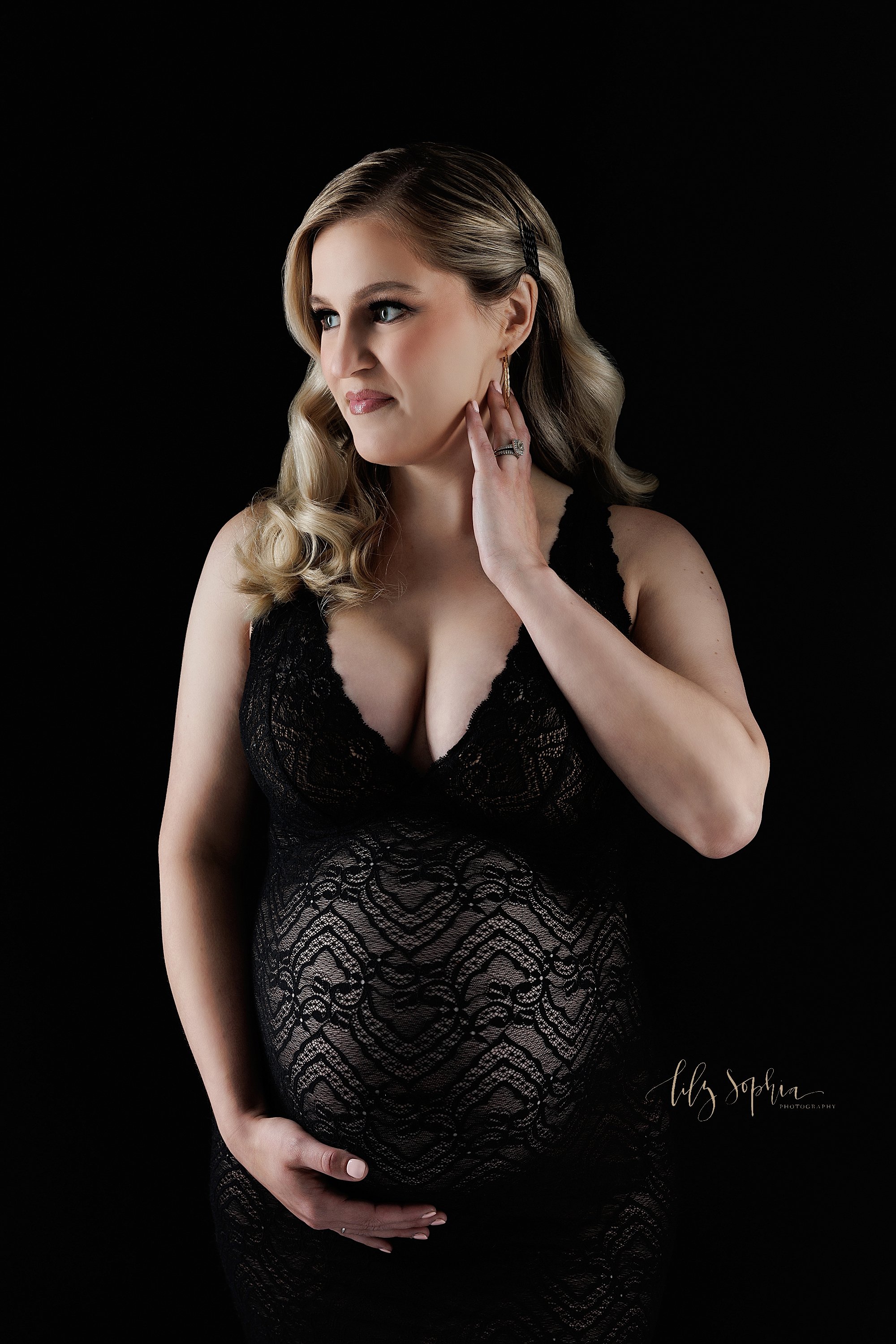  Dramatic maternity shoot in a modern maternity style as the pregnant mother wears a black plunging V-neck lace gown with the mother looking over her right shoulder as she holds the base of her belly with her right hand and places her left hand on he