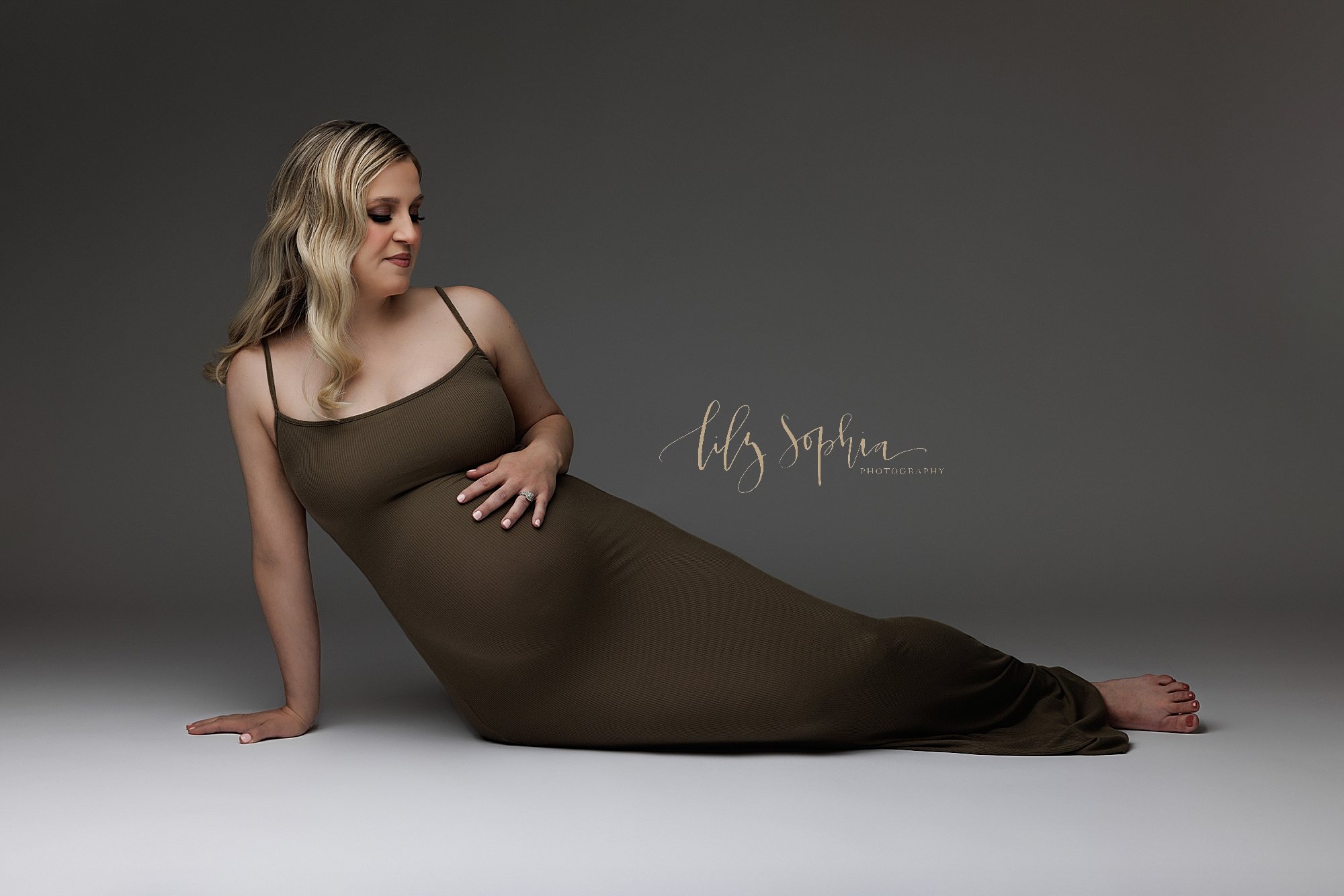  Maternity portrait of a pregnant mother wearing a spaghetti strap jersey knit gown as she lies on her right hip with her right hand supporting her body and her legs to the left of her body and her left hand placed on top her belly as she looks to he