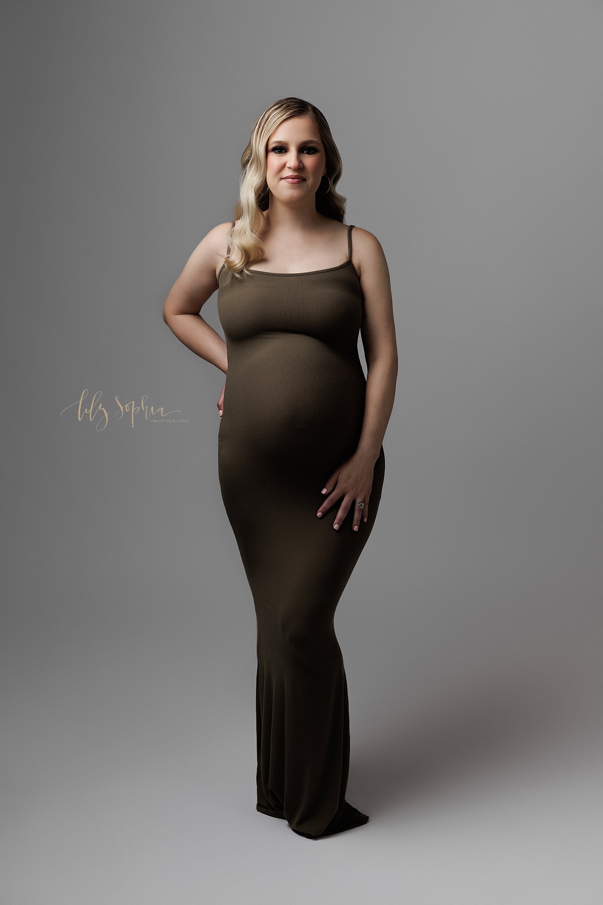  Modern maternity session with a pregnant mother as she stands in a spaghetti strap full length jersey knit gown with her right hand on her hip and her left hand on her thigh in a photography studio near Midtown in Atlanta, Georgia. 