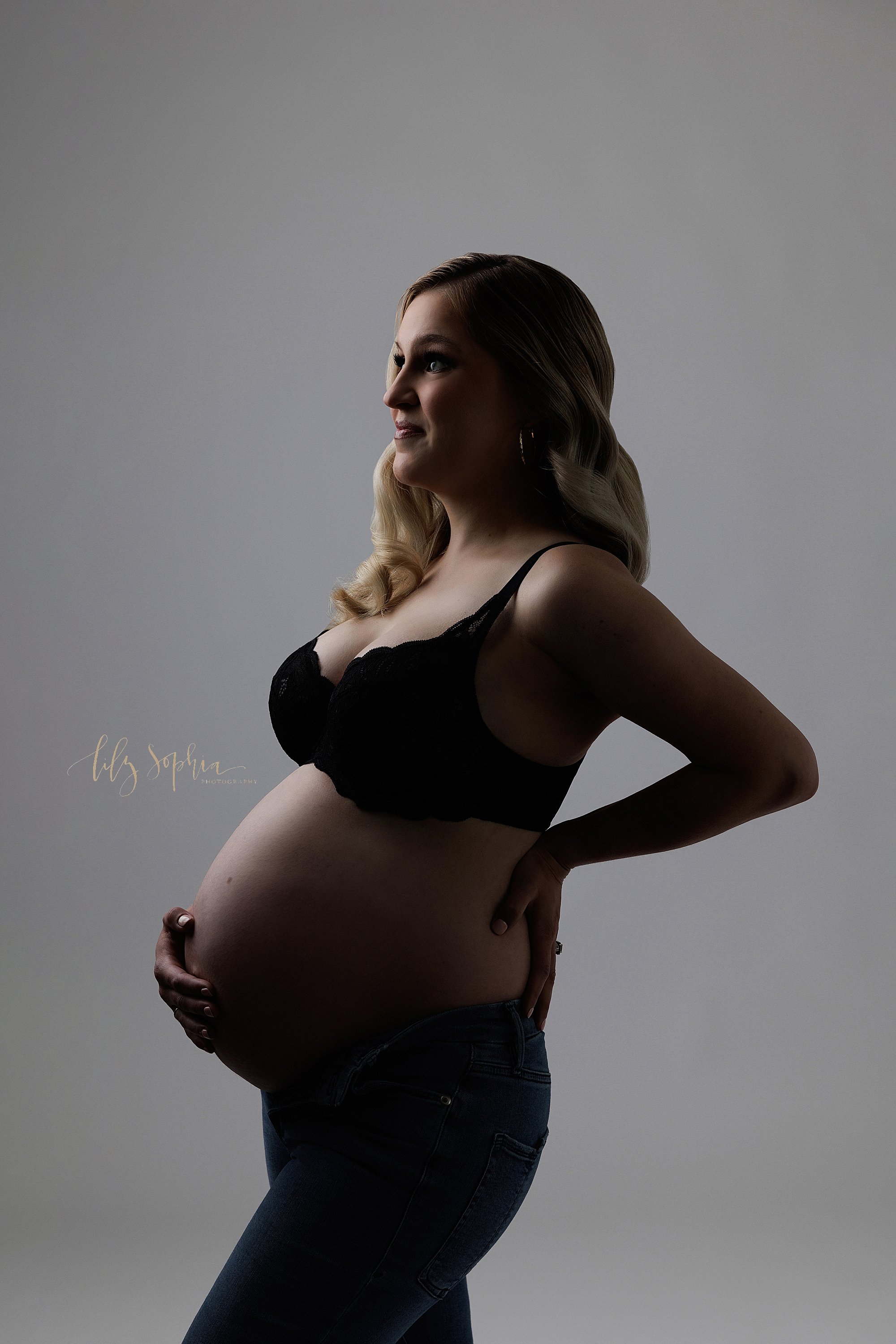  Modern maternity portrait taken in a photo studio of a pregnant mother wearing a black lace bra and an unzipped pair of blue jeans as she stands with her profile highlighted placing her right hand on her bare belly and her left hand on her hip taken