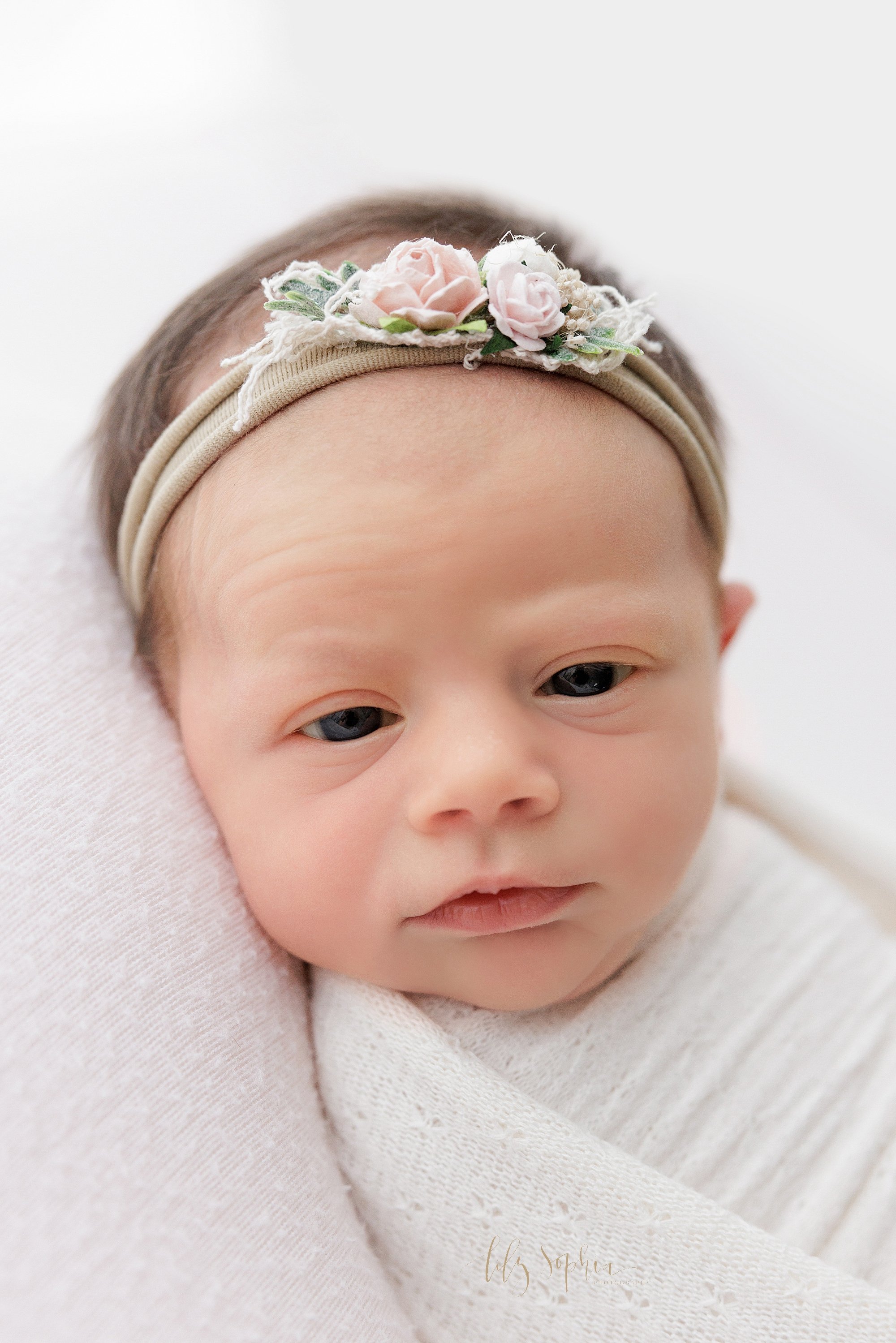  Close-up photo of an awake newborn baby girl as she lies on her back wrapped to her chin in a soft white blanket and wearing a headband in her hair as she looks over her right shoulder taken in a natural light studio near Poncey Highlands in Atlanta