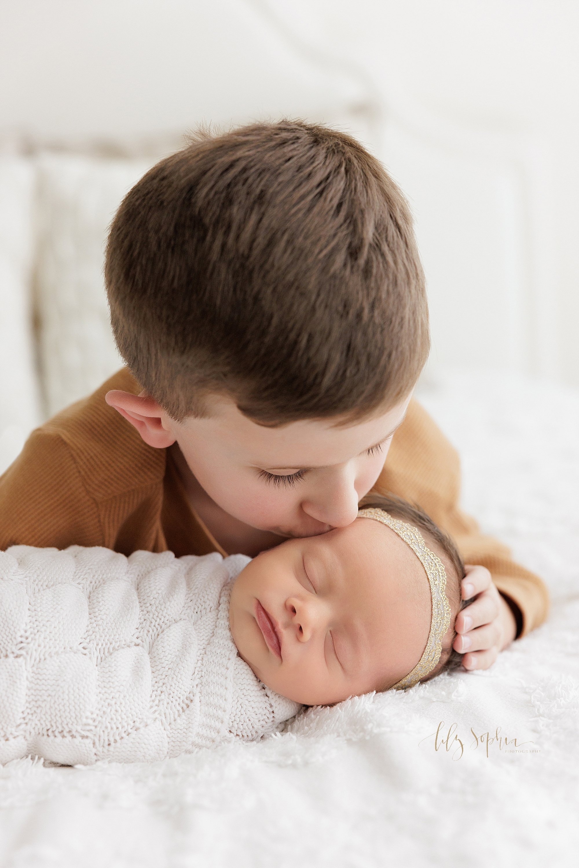  Close-up newborn sibling portrait of a big brother lying on his stomach on a bed as he leans over his peacefully sleeping baby sister as he kisses her on her forehead and gently places his right hand on her head taken in a photography studio near Vi