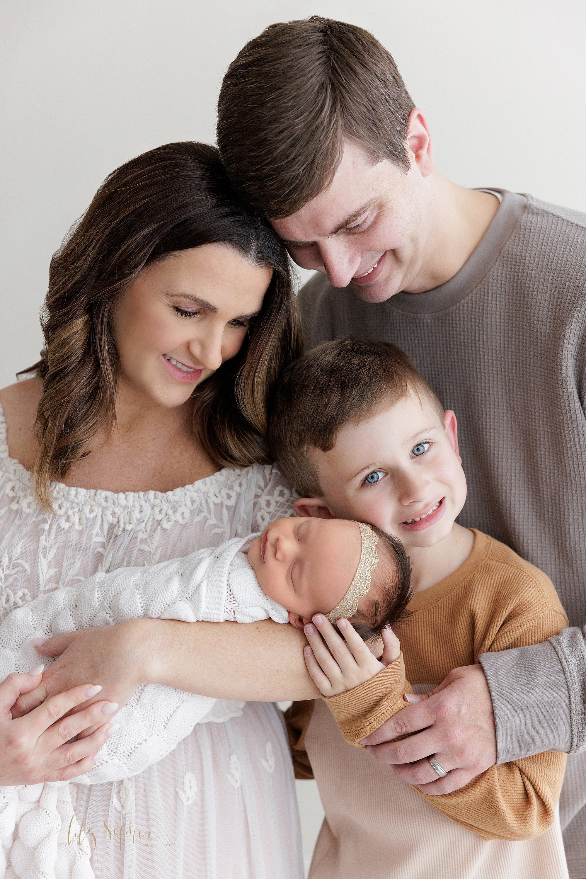  Family newborn photo with mom holding her peacefully sleeping newborn baby girl in her arms as her son places his cheek  against his sister’s head and dad stands behind his son as mom and dad look at their daughter taken in natural light in a photog