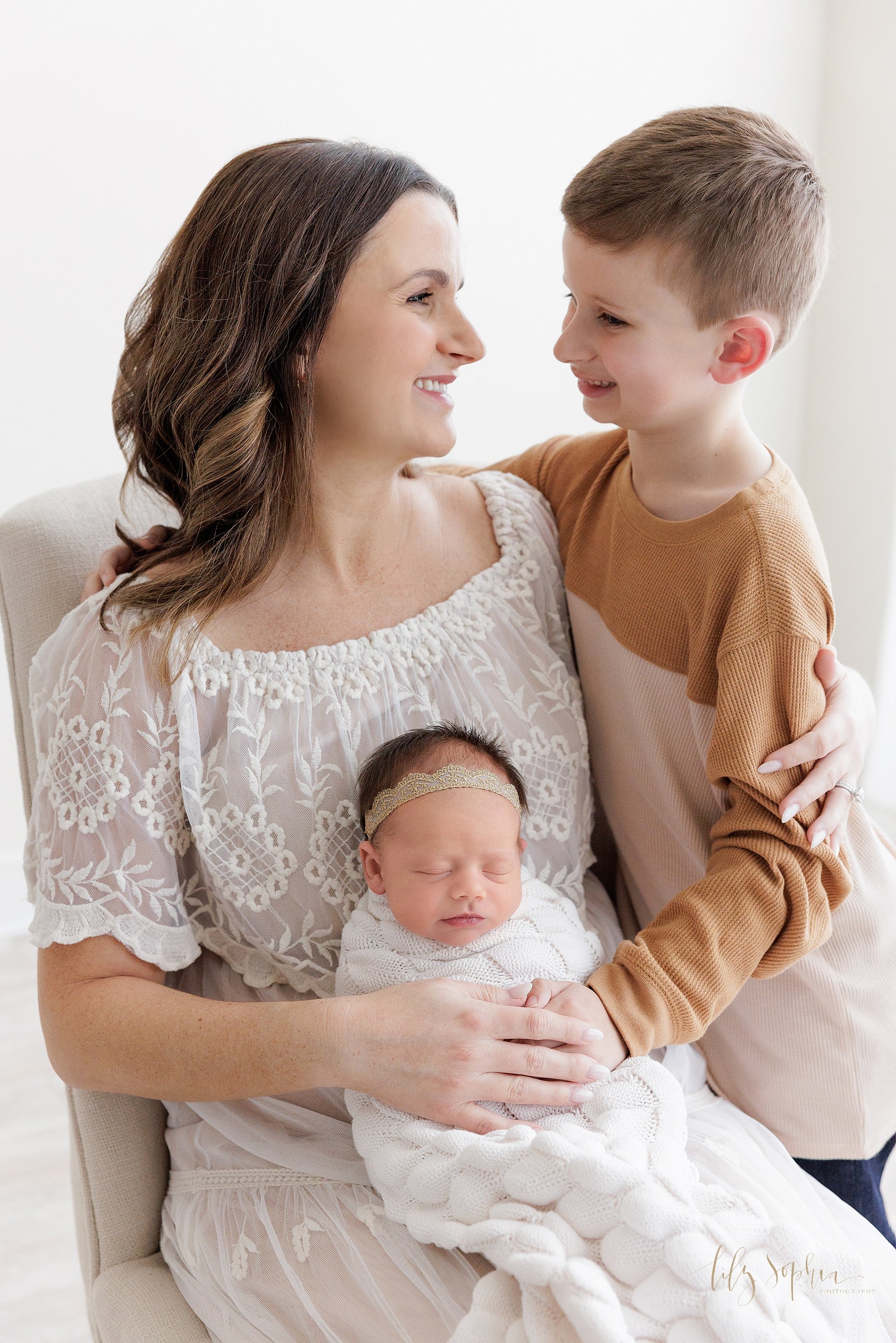  Maternity mother and children portrait as mom sits in a chair with her newborn baby girl on her lap and her young son stands to her left side with his arms around his mother’s back as mom and son talk to one another with mom  placing her left hand o