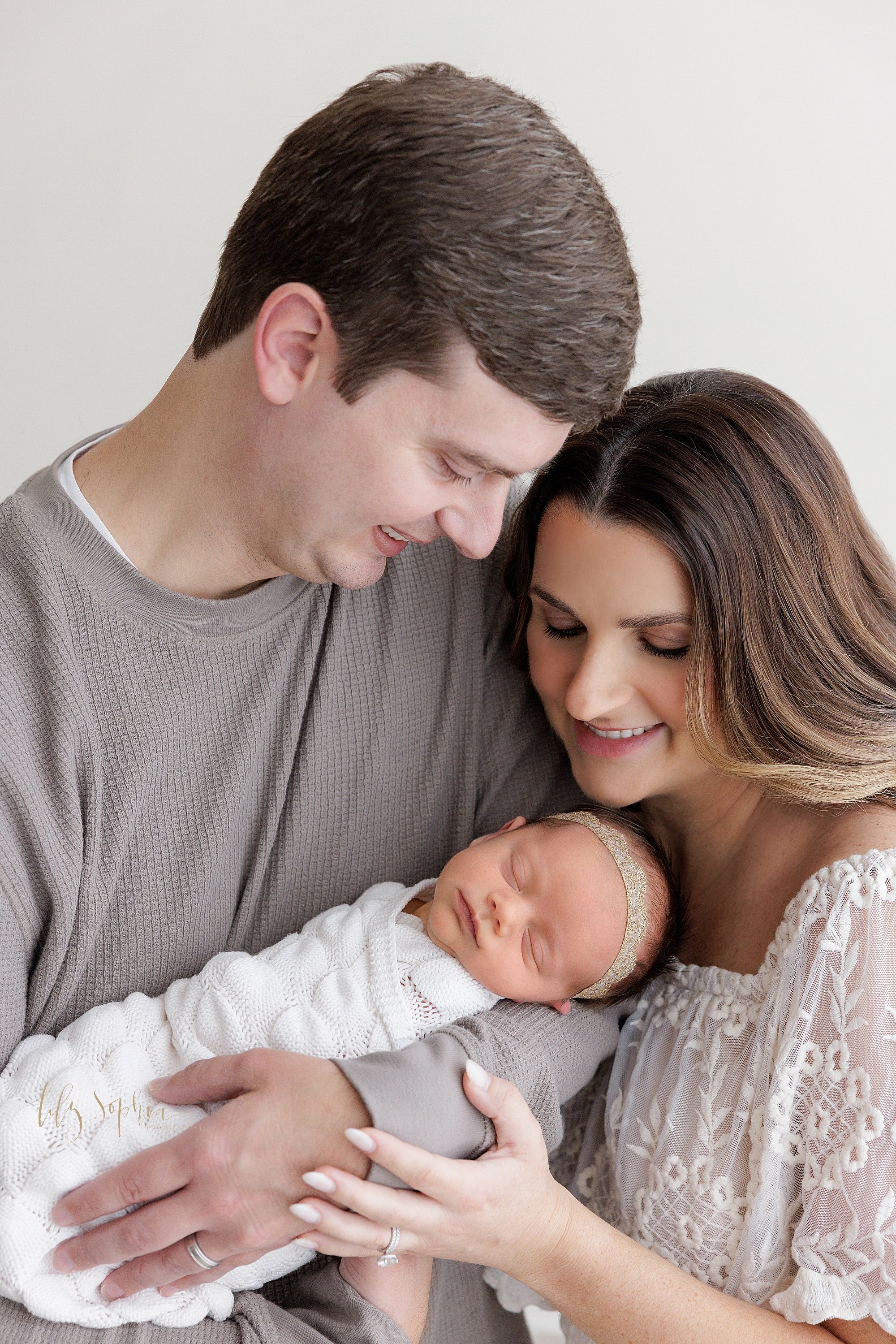  Newborn portrait of a father cradling his newborn baby girl  in his arms with his wife standing on his left side and placing her left hand on his right hand while the couple admires their newborn daughter taken near Brookhaven in Atlanta in a natura