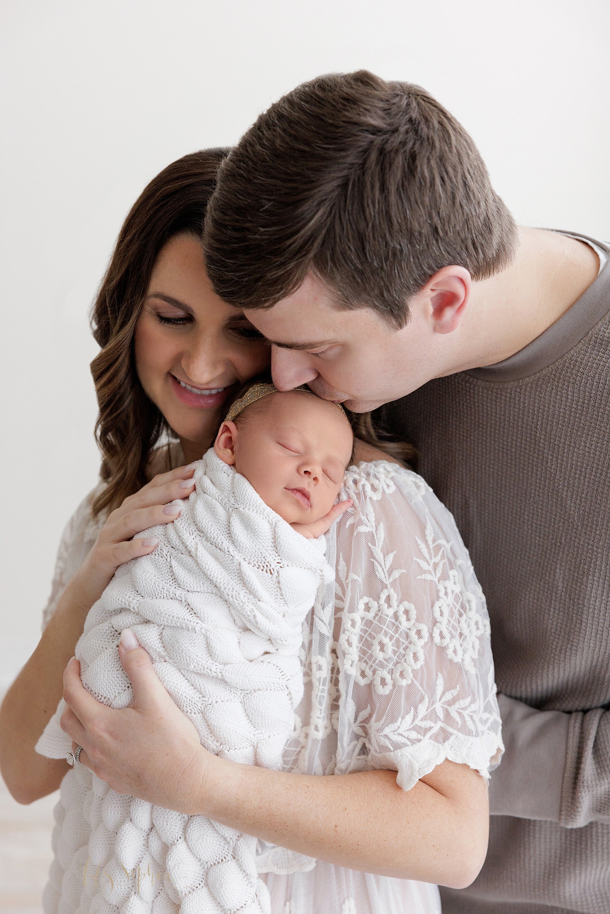  Newborn picture of a sleeping newborn baby girl being held against her mother’s left shoulder her husband stands behind her and bends over her shoulder to kiss their daughter/s forehead taken in natural light in a photography studio near Ansley Park