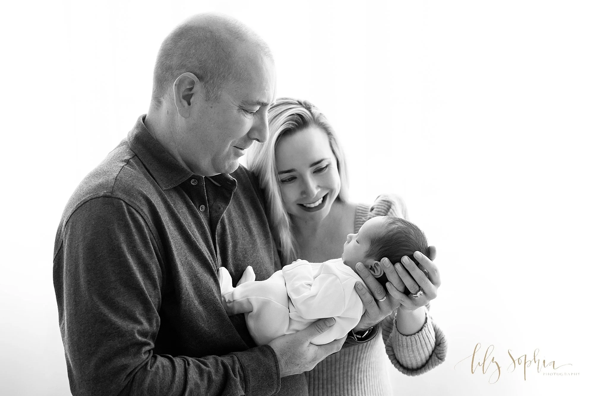  The wonder and admiration of a newborn baby boy is captured in a black and white photo of a father holding his newborn son in his hands as the mother places her left hand on her son's head as she stands next to her husband in a photography studio ne