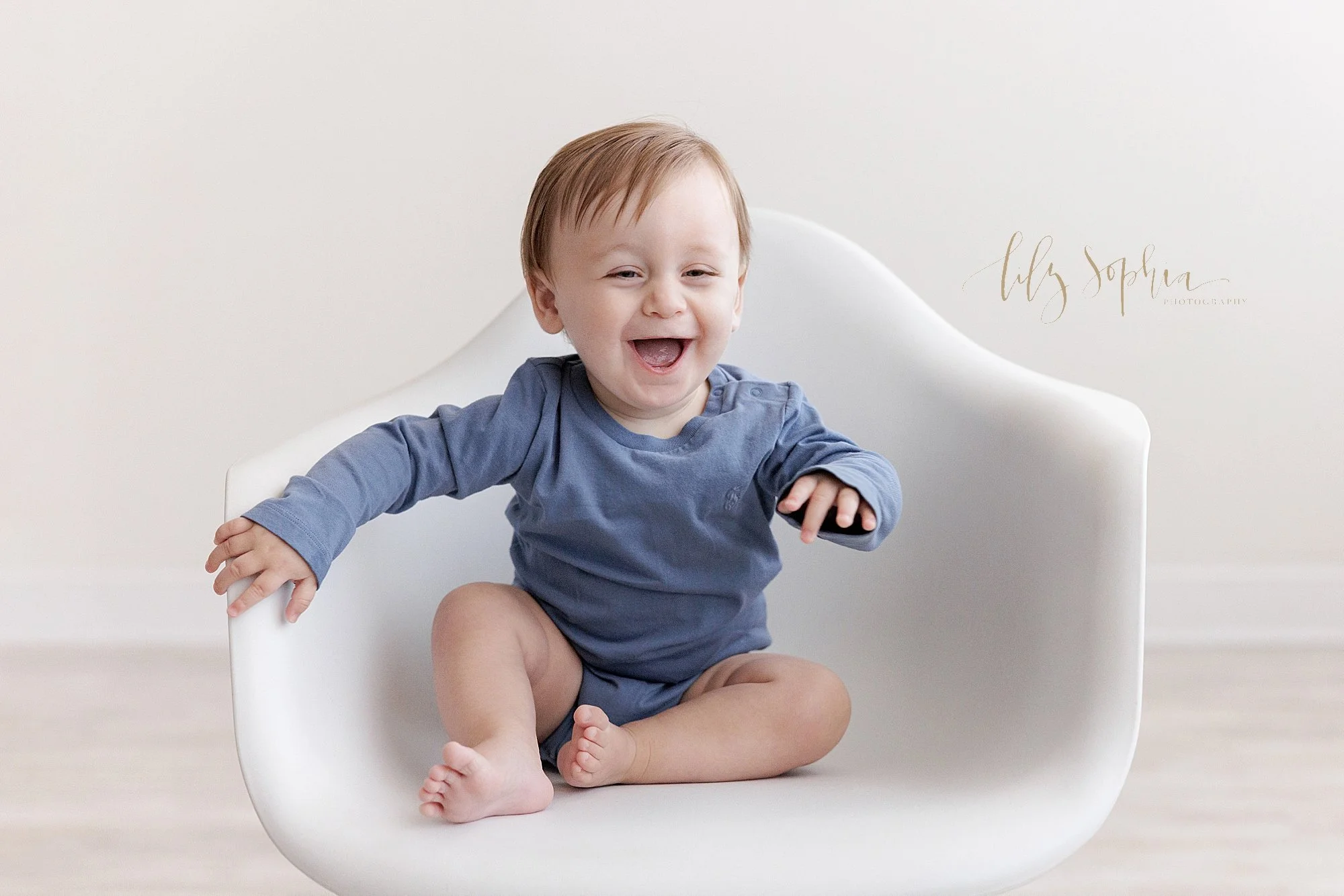  A proud and happy one year old boy sits in a white molded chair next to a window streaming natural light into a photography studio for a first birthday session near Ansley Park in Atlanta. 