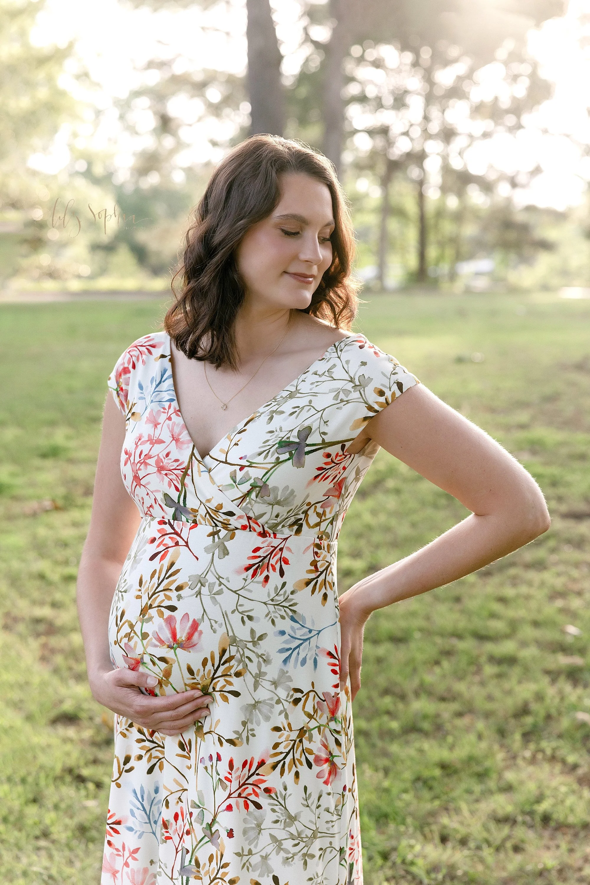 Maternity photo session of an expectant mother as she stands at sunset in an Atlanta park with her right hand on her hip and her left hand holding the base of her womb.