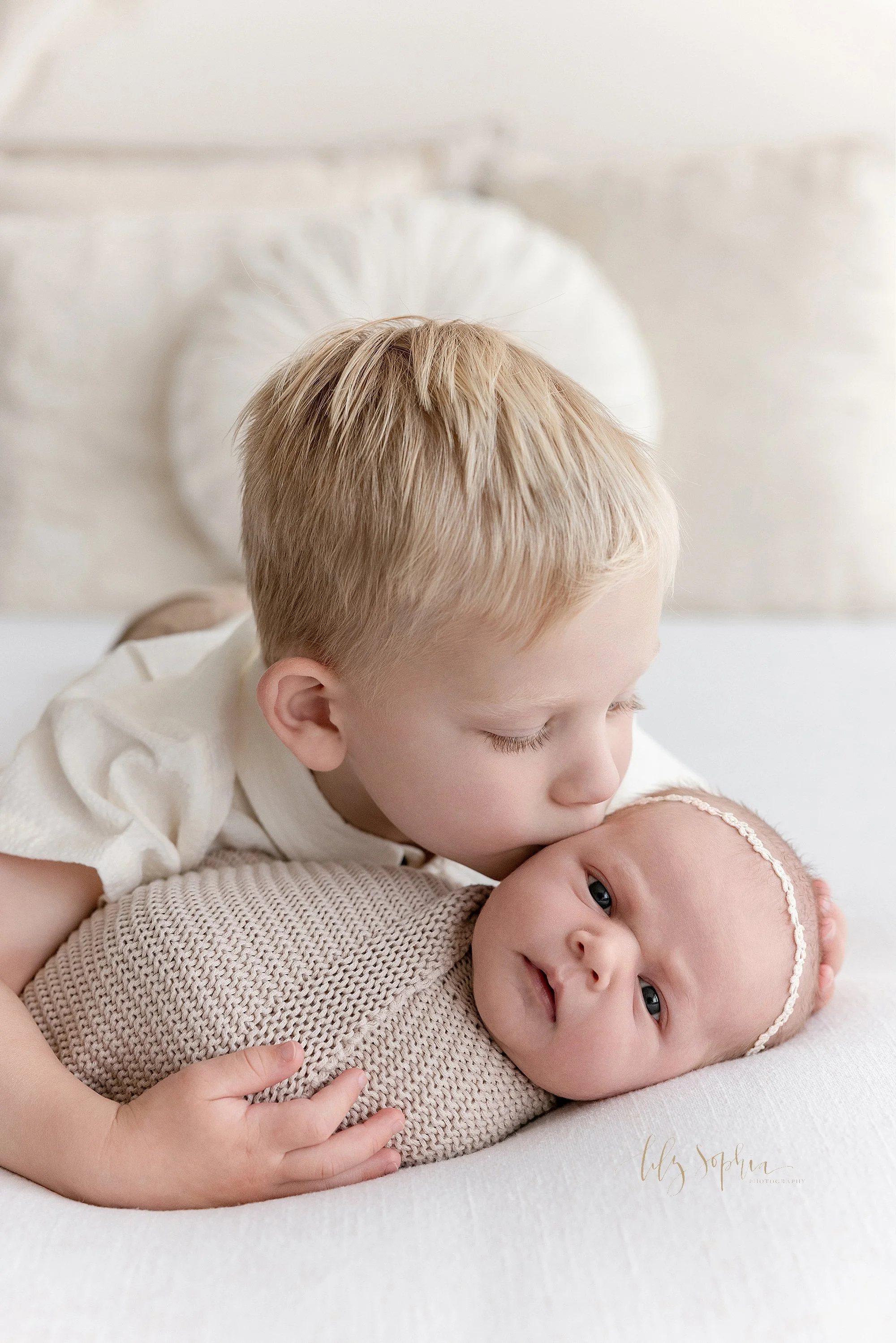  Close-up newborn photo of an awake newborn baby girl swaddled in a knitted blanket lying on her back as her older brother lies on his stomach and kisses the right side of her head is taken near Morningside in Atlanta using natural light in a photogr