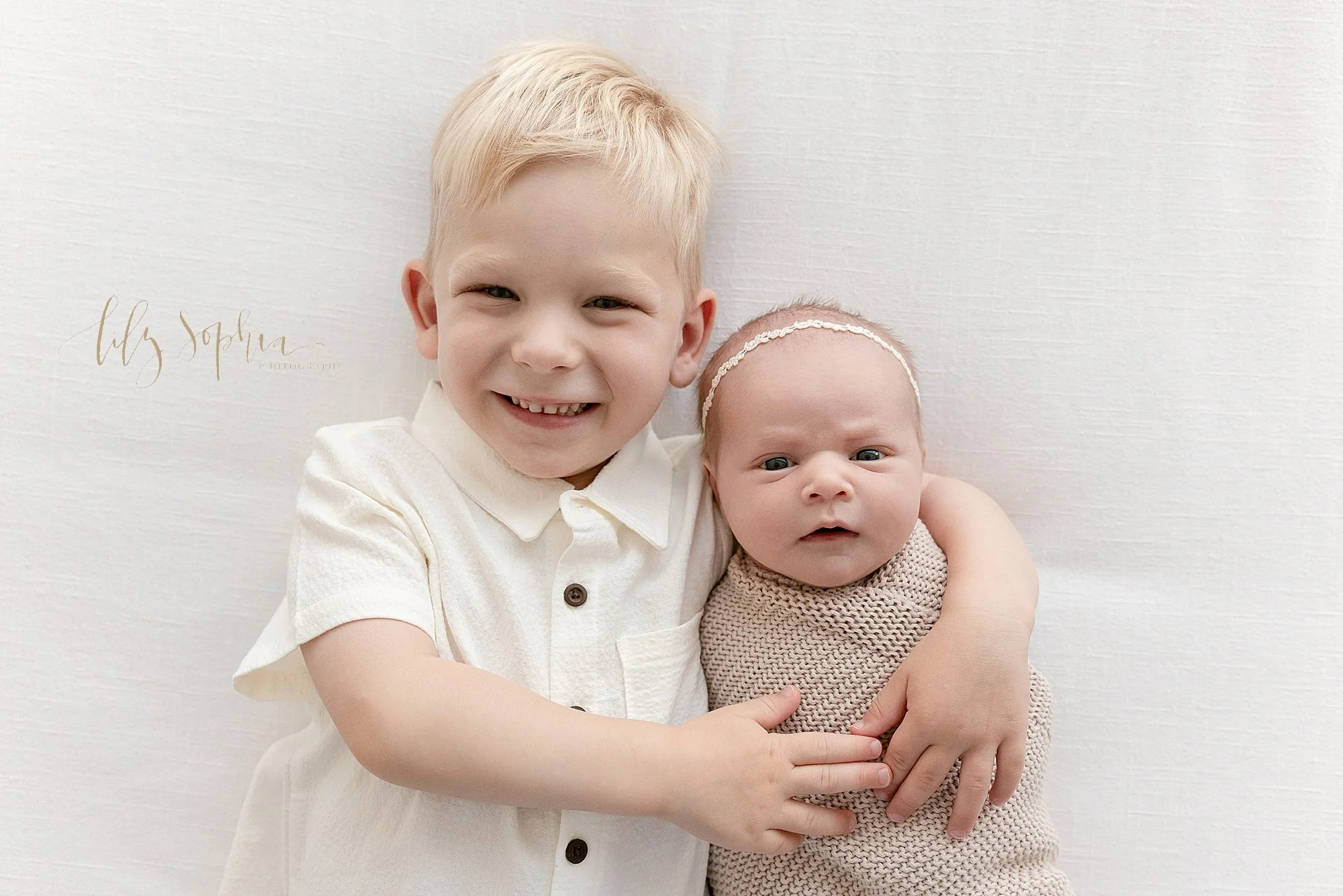  A proud older brother lies on his back and wraps his arms around his awake newborn baby sister in a photography studio near Vinings in Atlanta that uses natural light. 