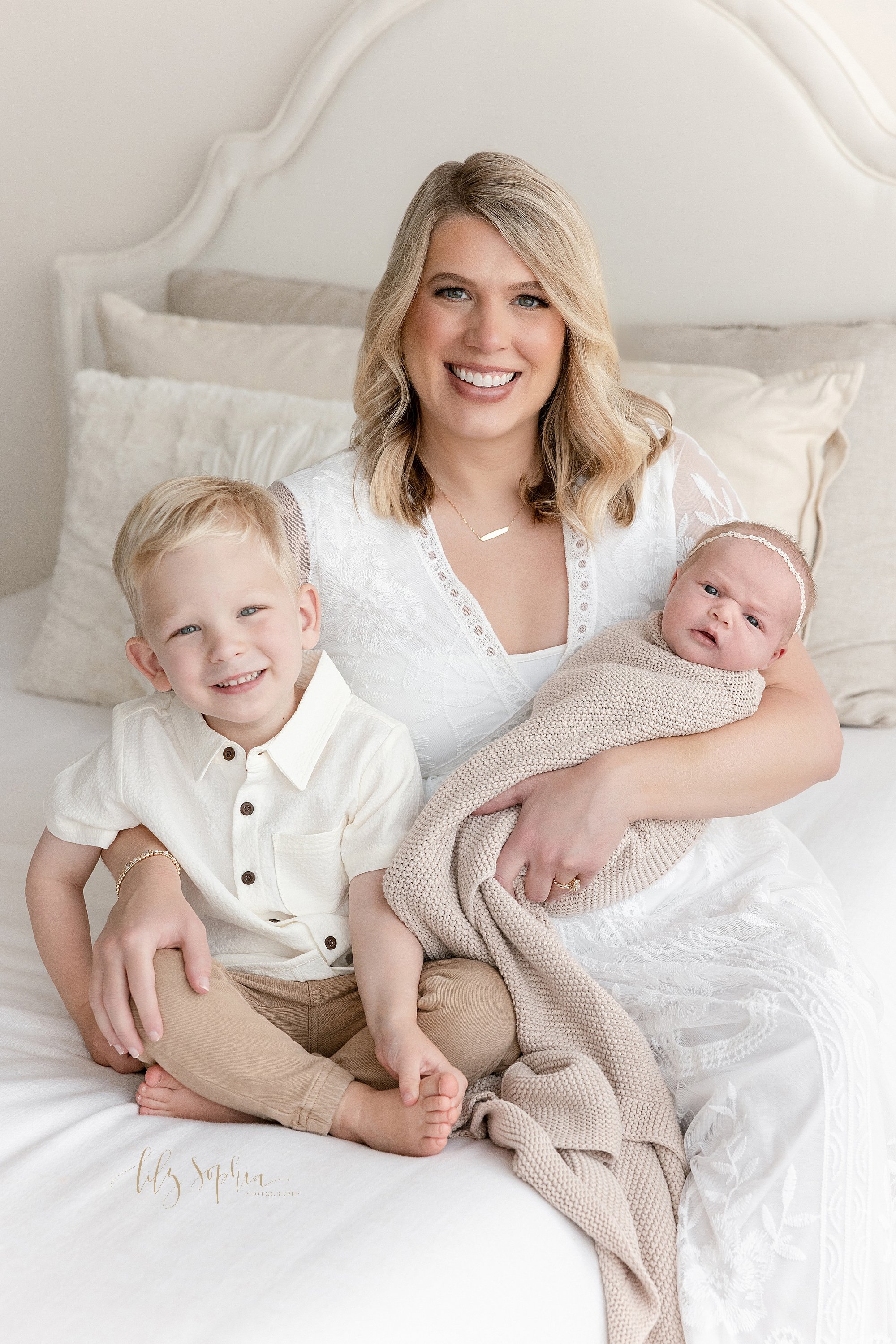  Newborn family portrait of a mother sitting on a bed holding her newborn baby girl in her right arm as her young son sits next to her and she places her left hand on his knee taken in a photography studio near Alpharetta in Atlanta that uses natural