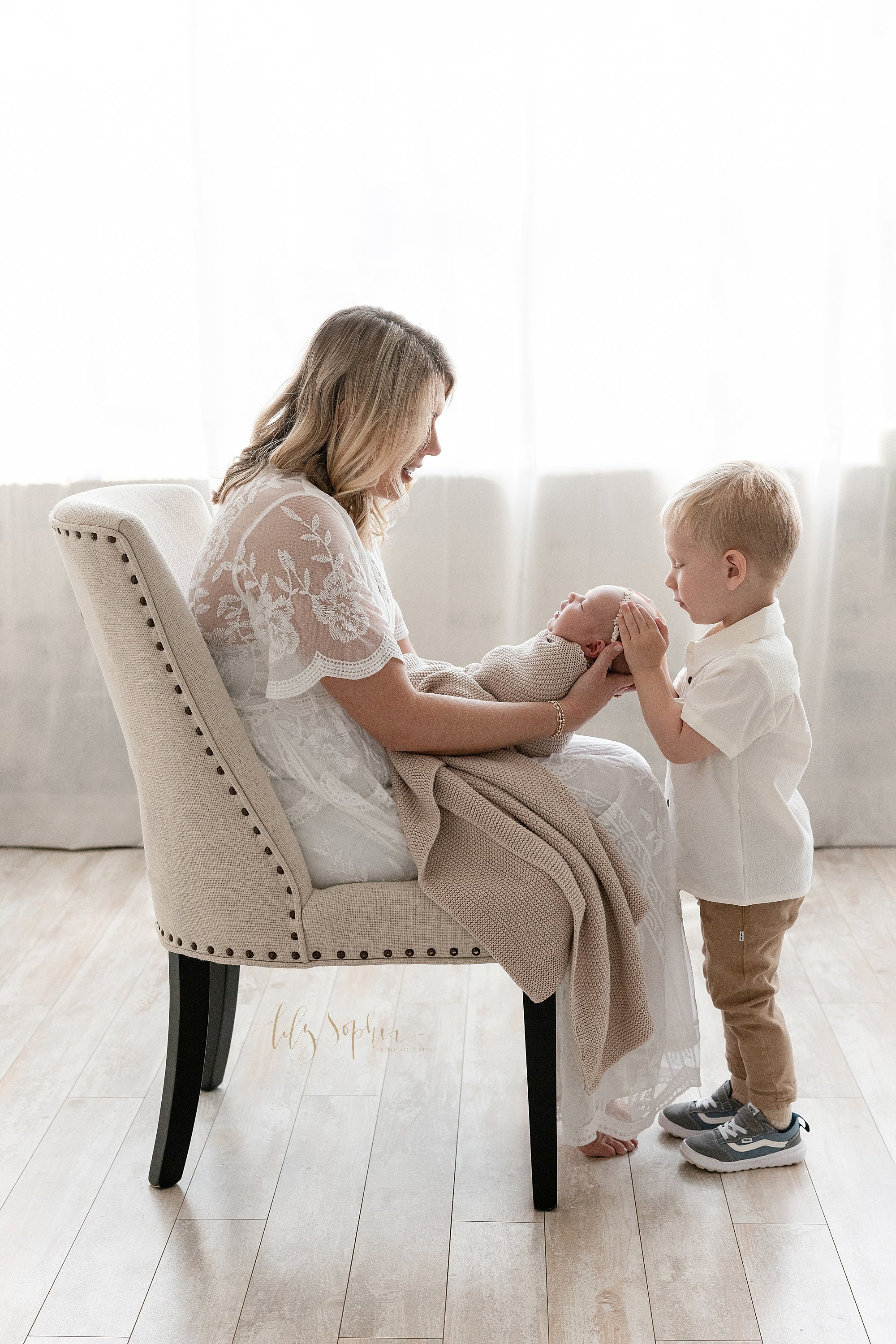  A mother sits in an upholstered chair as she holds her newborn baby girl in her hands and her young son stands facing her with his hands on his sister’s head while natural light streams into the photography studio from behind that is located in Ponc