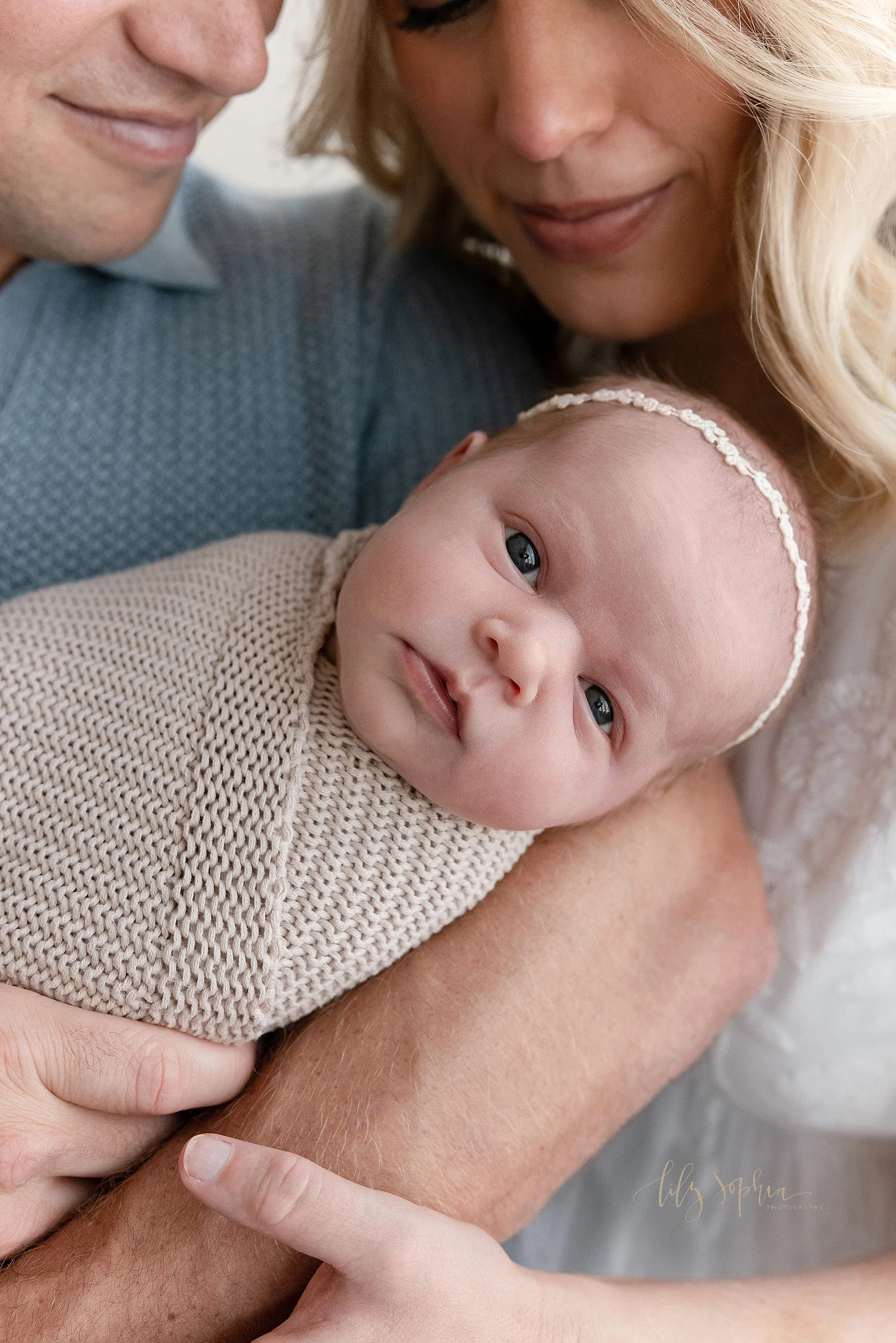  Close-up picture of an awake newborn baby girl as her father holds her and her mother looks on taken near Cumming in Atlanta in a natural light photography studio. 