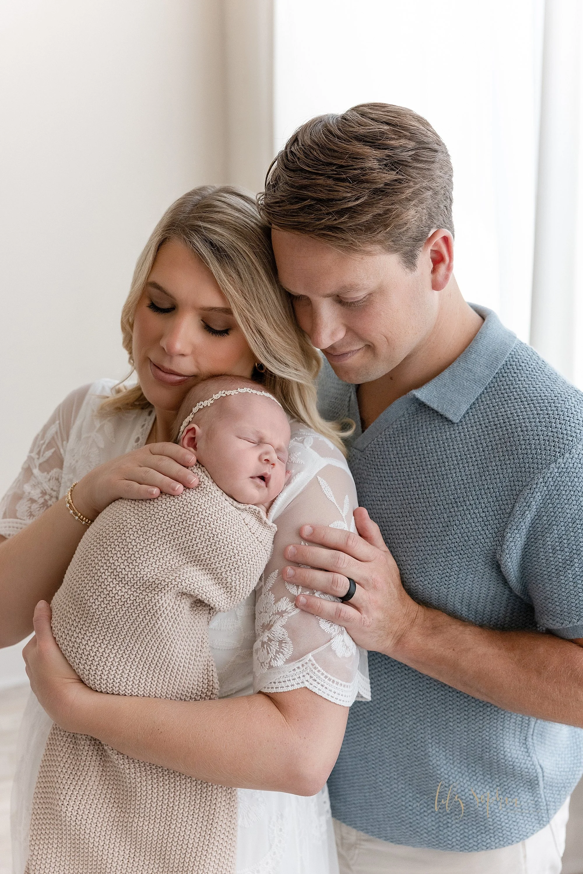  The treasured peaceful time is captured during this newborn photography shoot with mom holding her newborn baby girl against her left shoulder as dad stands behind her and places his hand on his wife’s left arm as the two stand in front of a window 