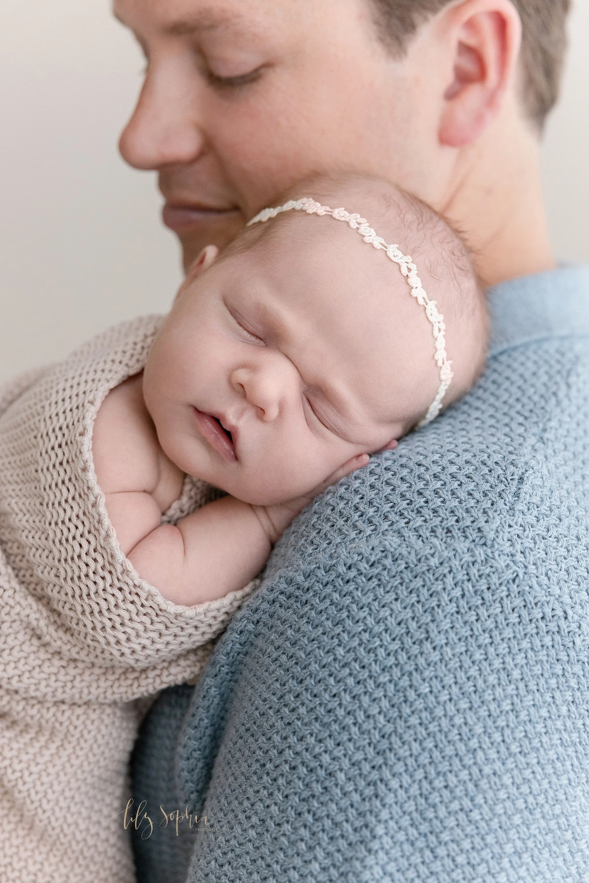  Close-up photo of a sleeping baby girl as she is held against her father’s left shoulder taken using natural light near Roswell in Atlanta in a photography studio for a newborn session. 