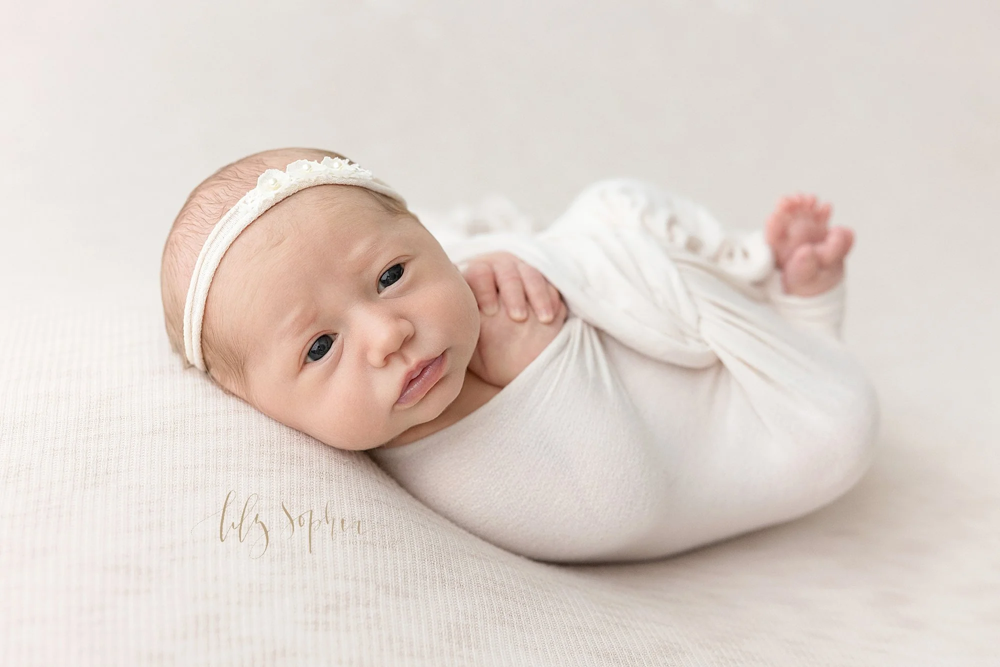  A wide awake newborn baby girl lies on her back wearing a stretchy flowered headband in her wispy hair as she is bundled in a stretchy swaddle with her hands folded beside her face and her feet sticking out taken near Brookhaven in Atlanta in a phot
