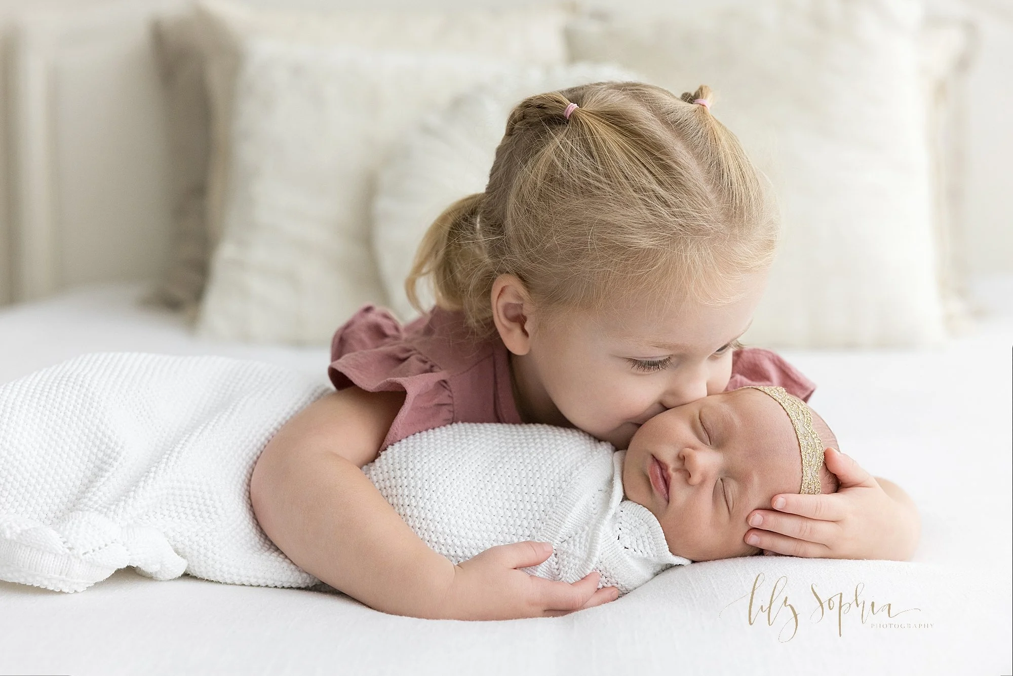  Sibling photo during a newborn photography session with an older sister lying on her stomach as she wraps her arms around her newborn baby sister and kisses her sister on her cheek taken in a studio that uses natural light near Vinings in Atlanta. 