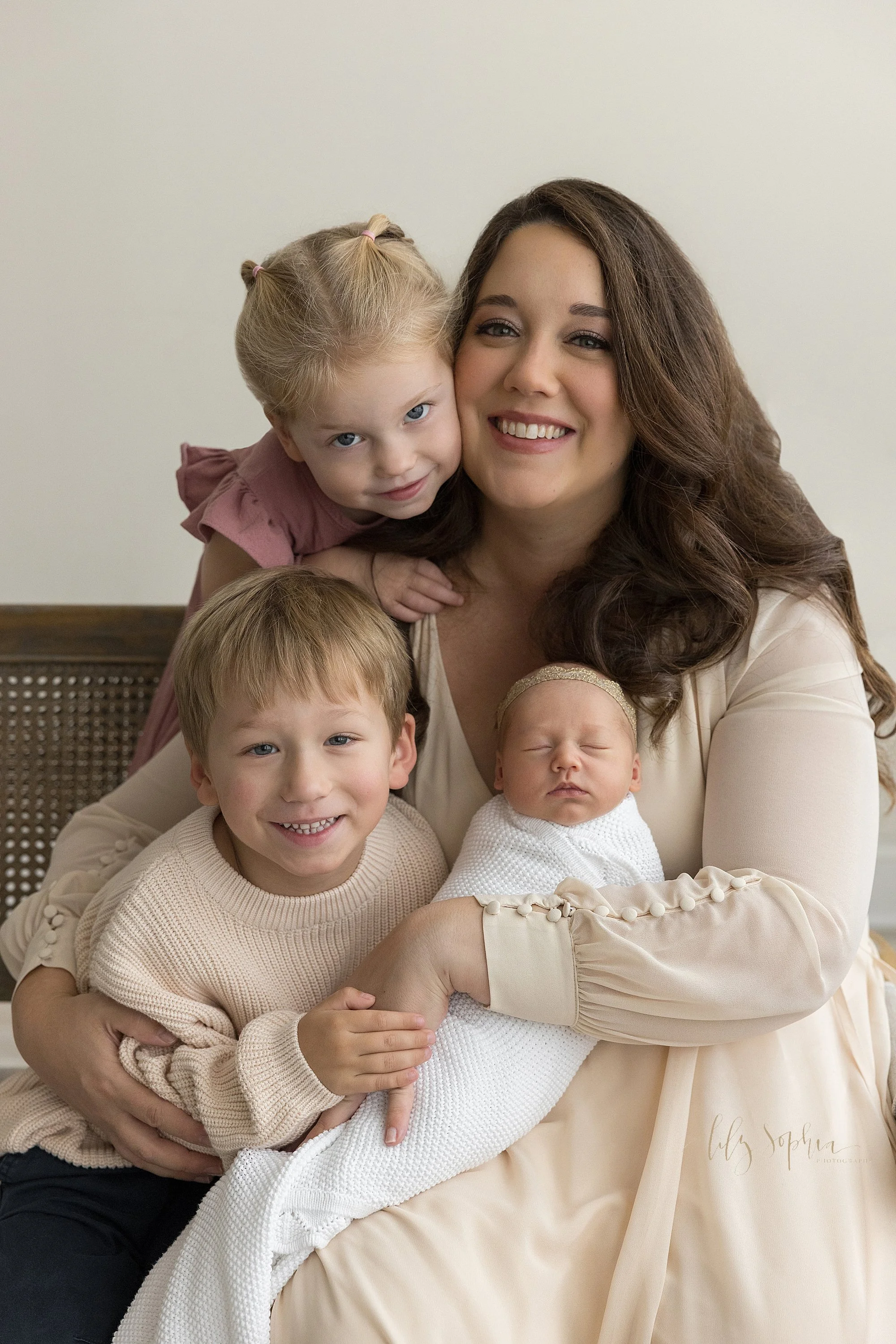  Newborn family photo taken during a first year collection photography session with mom holding her newborn baby girl on her lap and holding her with her left hand as her young son sits to mom’s right and her young daughter stands behind her on her r