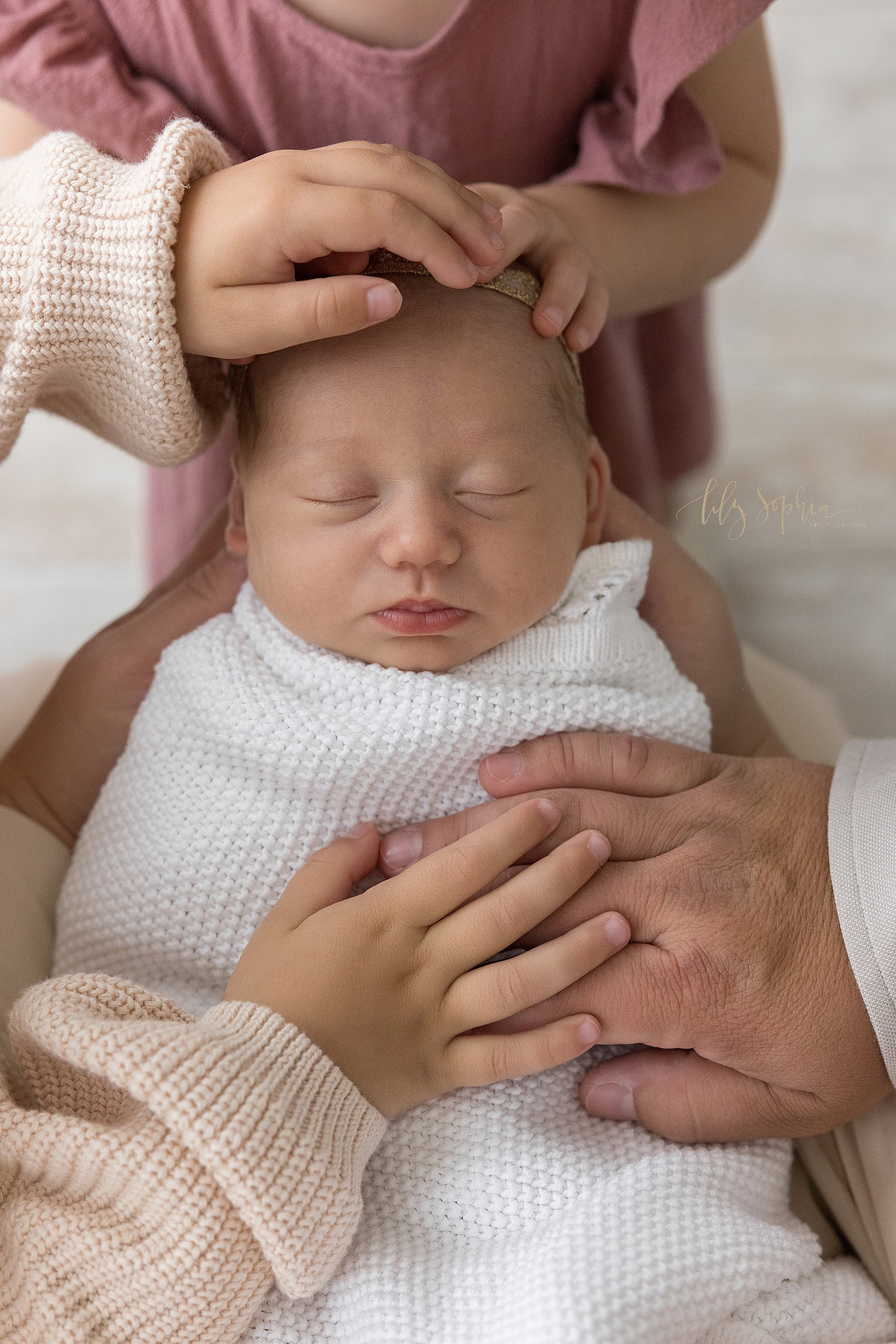  A newborn photo of a sleeping newborn baby girl as her mother holds her head in her hands and her siblings place their hands on their sister’s head and as her father places his hand on his daughter’s chest while his young son places his hand atop hi