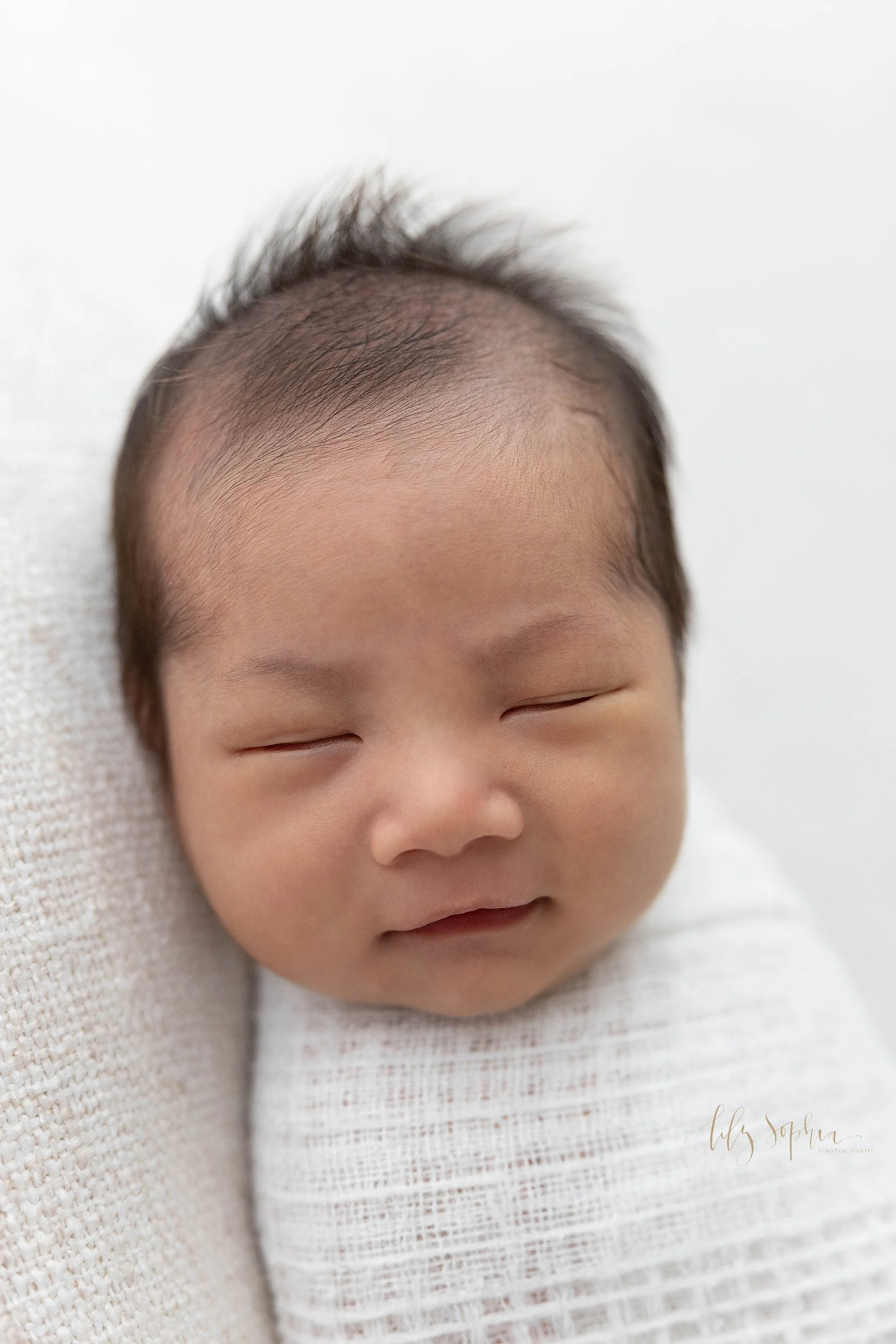  Close-up of a peacefully sleeping Asian newborn baby boy as he looks over his right shoulder while lying on his back taken in a photography studio that uses natural light near Buckhead in Atlanta. 