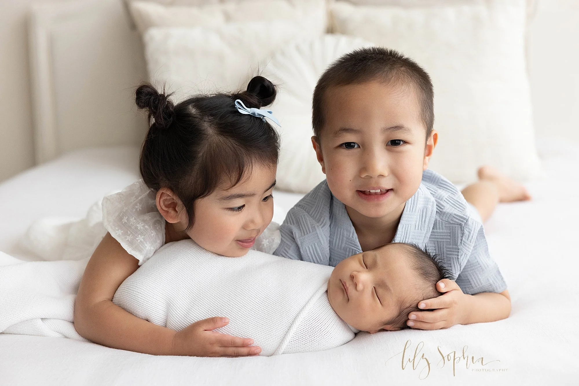  Newborn sibling photo of a newborn baby boy lying on his back swaddled to his chin in a soft white blanket as his older sister lies on her stomach and reaches to hold him with her right arm while placing her chin on his leg and looks at him as her o