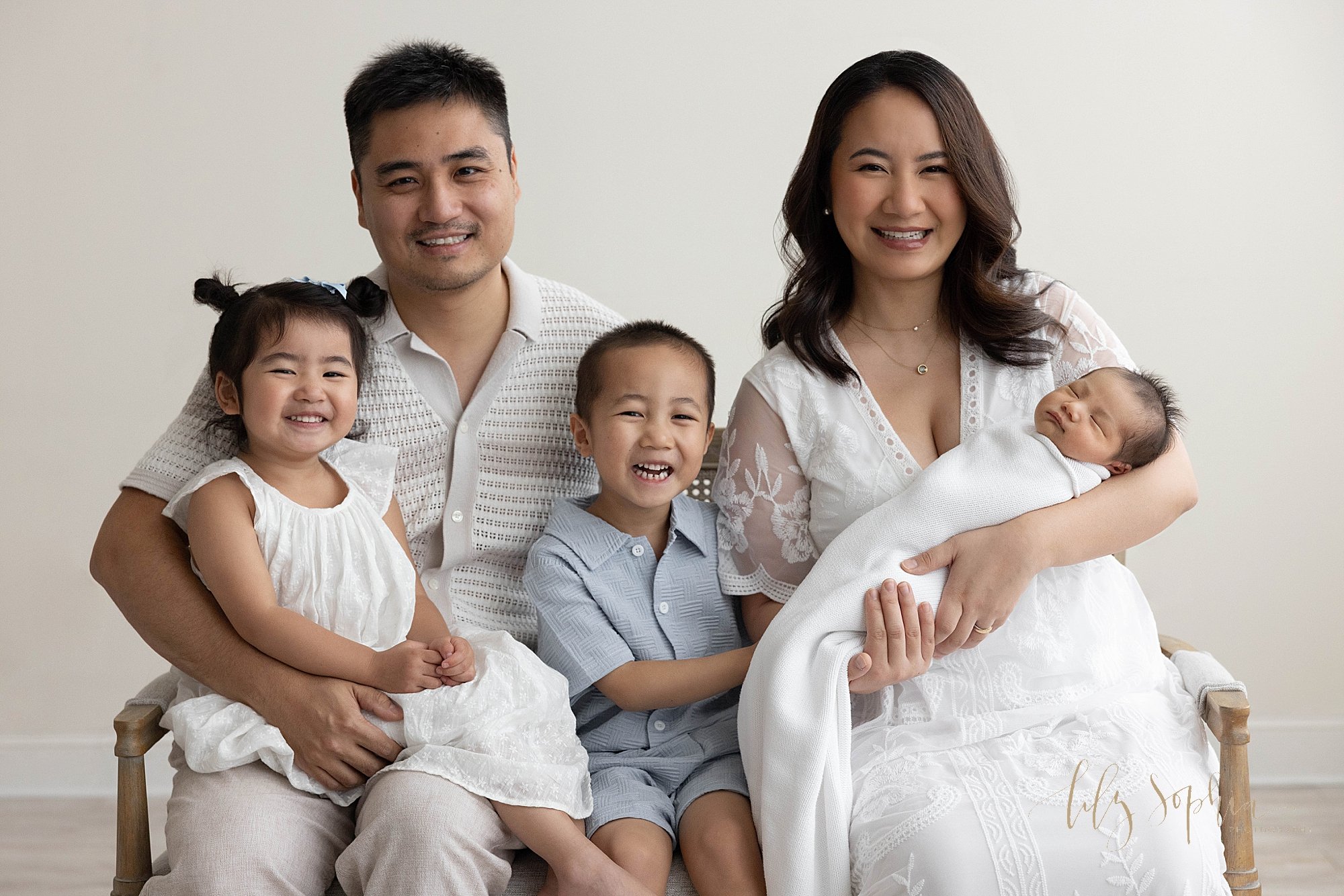  Newborn family photo with an Asian mom holding her newborn baby boy as her young son sits between her and her husband who is holding their young daughter on his lap taken near Smyrna in Atlanta in a natural light photography studio. 