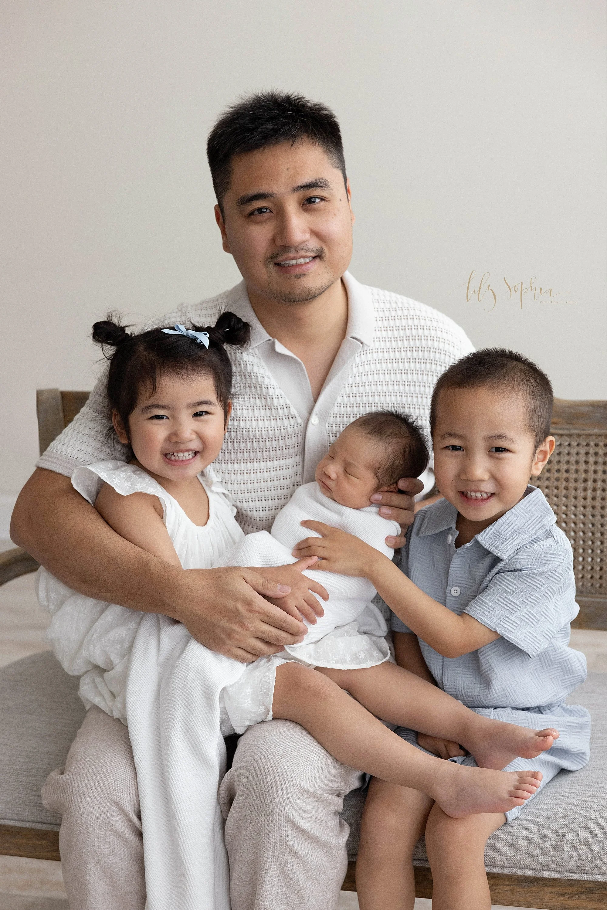  Newborn family father and children picture as the dad sits on a wooden bench with his young daughter on his right knee, his young son on his left knee and his newborn baby boy between them as he holds his baby boy’s head with his left hand taken in 