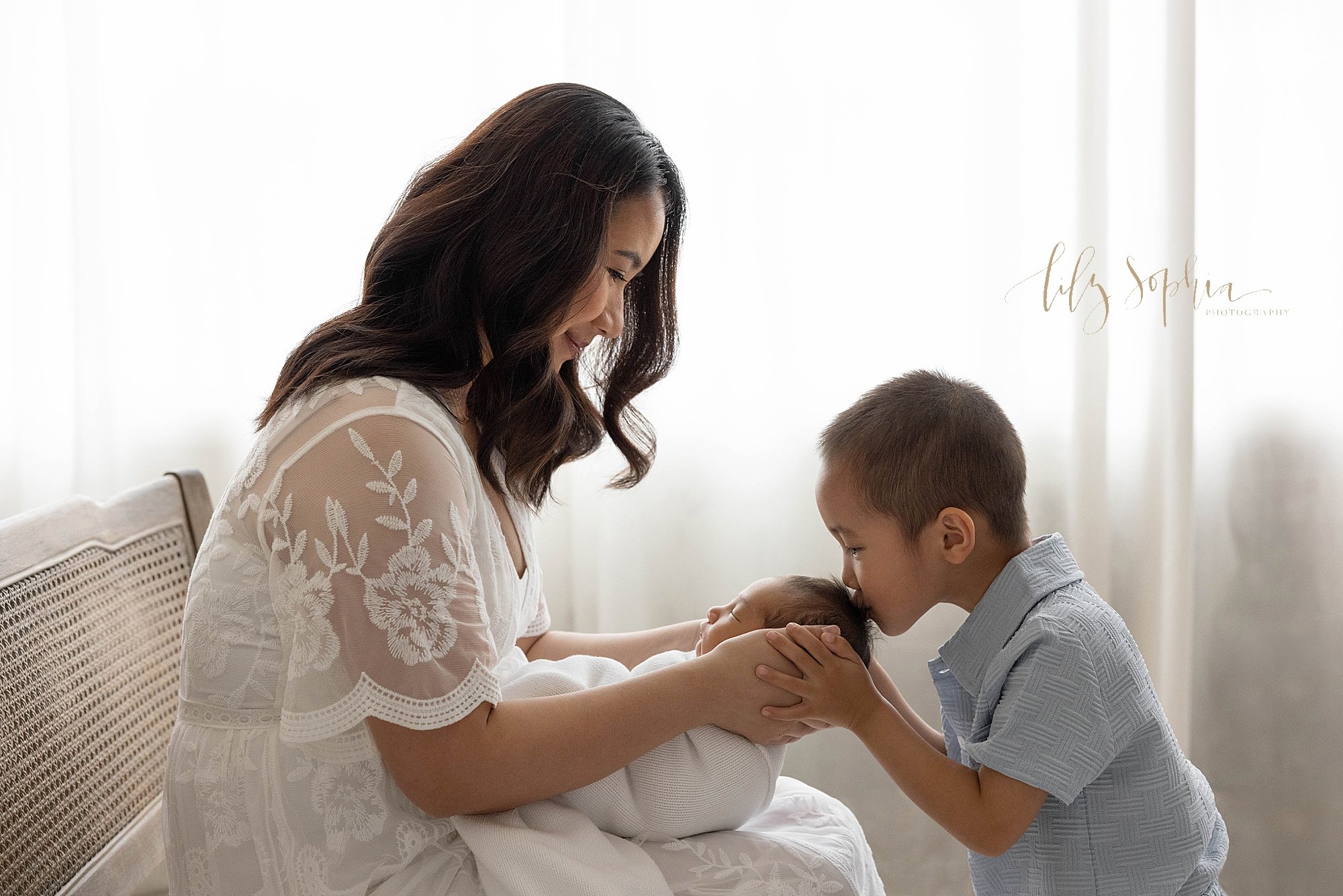  The love of a big brother for his newborn baby brother is captured during this newborn photography session as mom sits with her newborn baby boy on her lap as she holds his head while her young son  faces her and places his hands on hers and kisses 