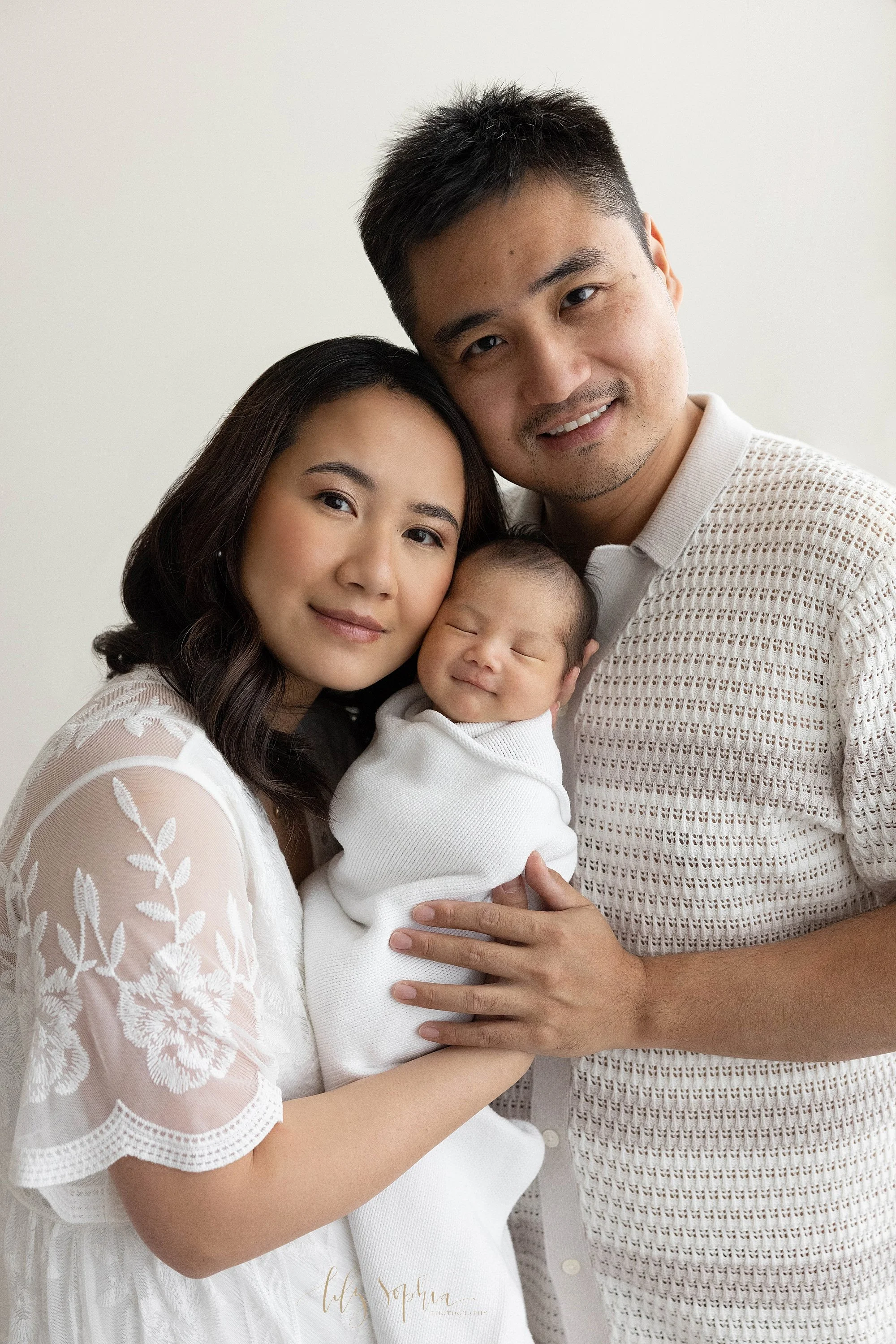  Family newborn portrait of a mom holding her smiling newborn baby boy against her left cheek as her husband faces her and leans his head against his wife’s and places his right hand on his wife’s taken near Kirkwood in Atlanta in a photography studi