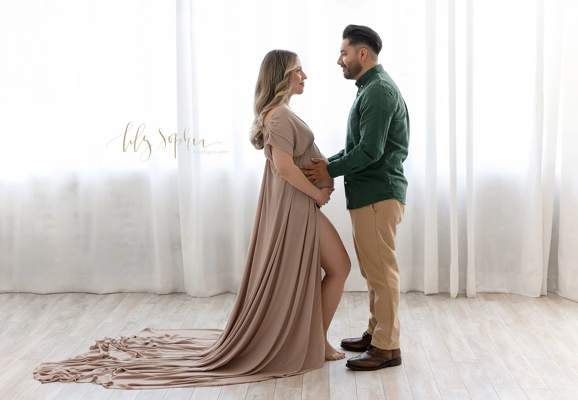  Maternity portrait of an expectant couple as they stand in front of a window streaming natural light into a photography studio located near Oakhurst in Atlanta with the husband facing the wife as he places his hands on their child in utero and she h