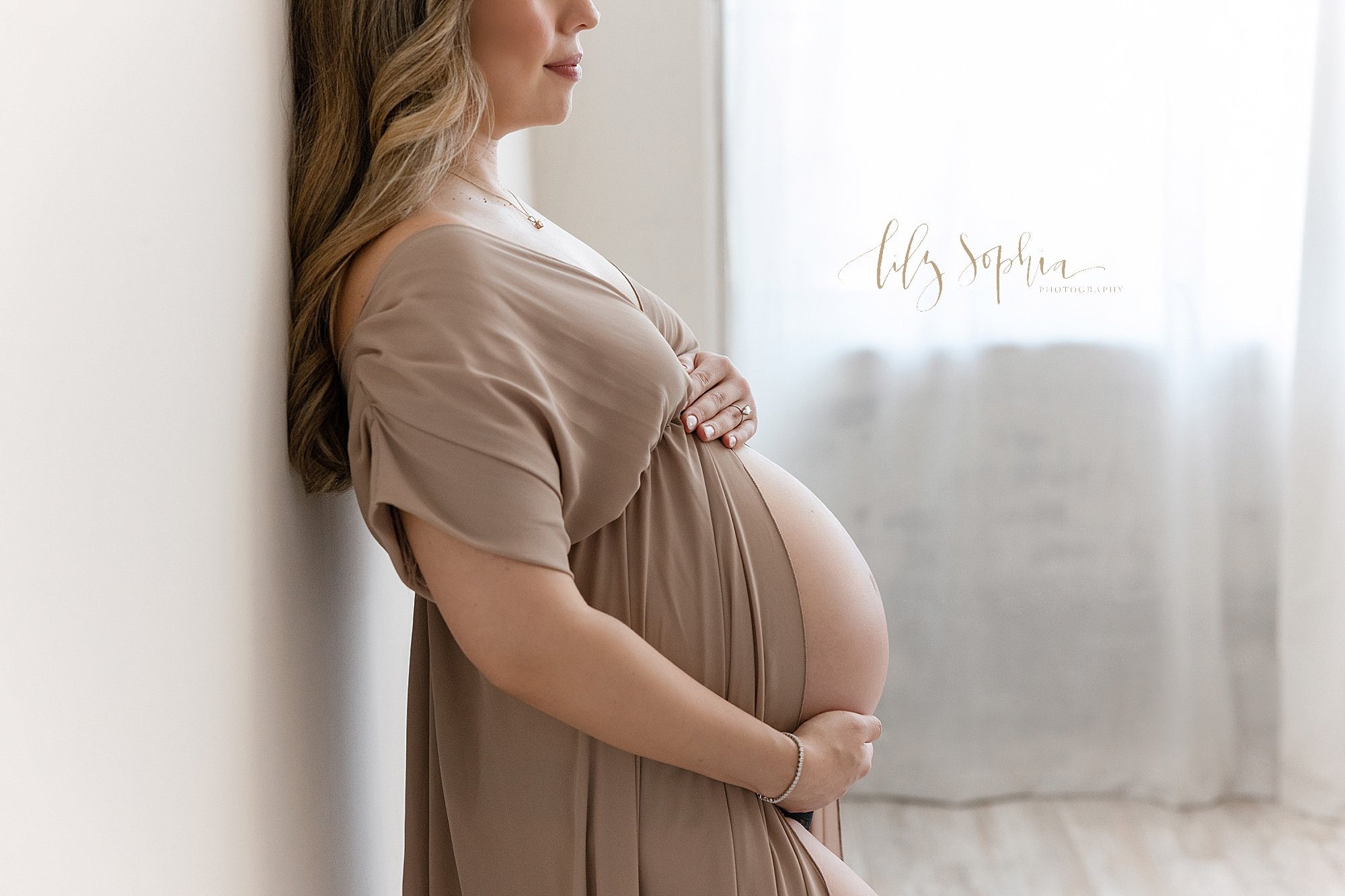  Maternity portrait of an expectant mother with her back against a wall wearing a split front jersey gown to bare her womb while she frames her belly with a window streaming natural light behind her taken in Ponce City Market in Atlanta, Georgia. 