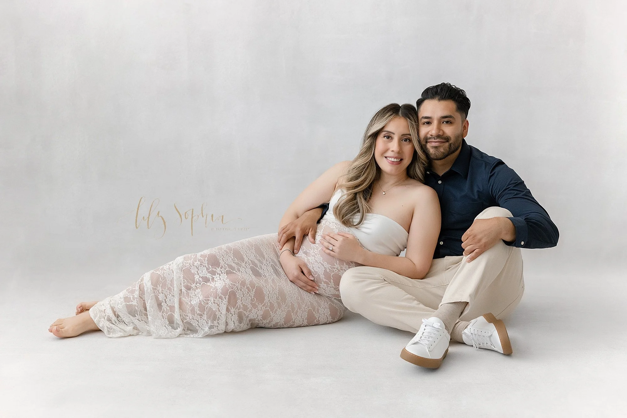  Maternity photo session in a natural light studio captures the expectant couple as the husband sits on the floor with his ankles crossed and holds his wife as she leans against him while framing her womb taken near Sandy Springs in Atlanta, Georgia.