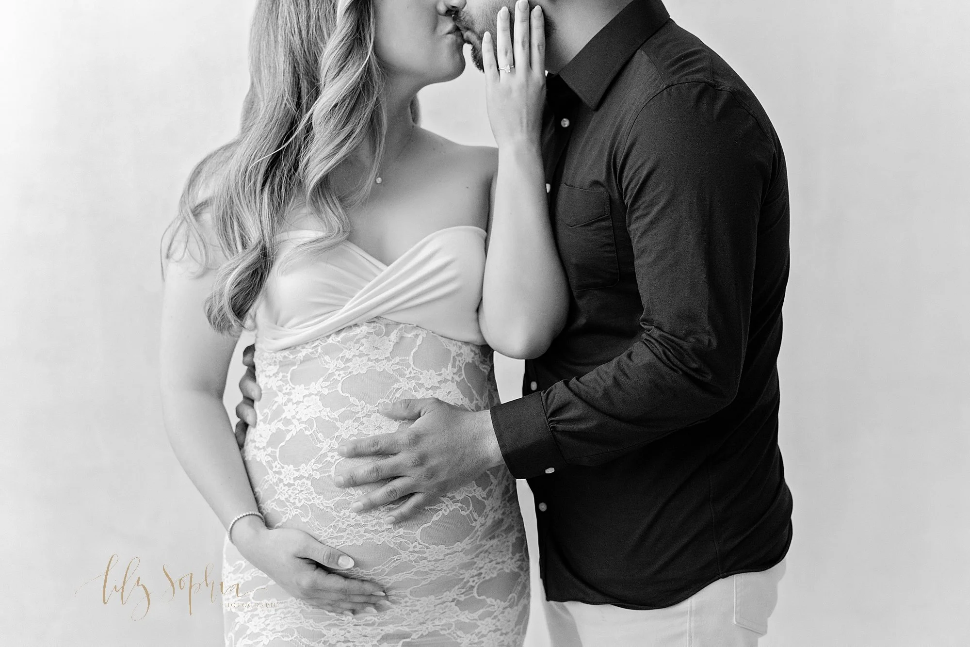  Black and white photo of a pregnant mother standing with her right hand holding the base of her womb as she grabs her husband’s right cheek and pulls him to kiss her while he places his right hand on their child in utero and his left hand around her