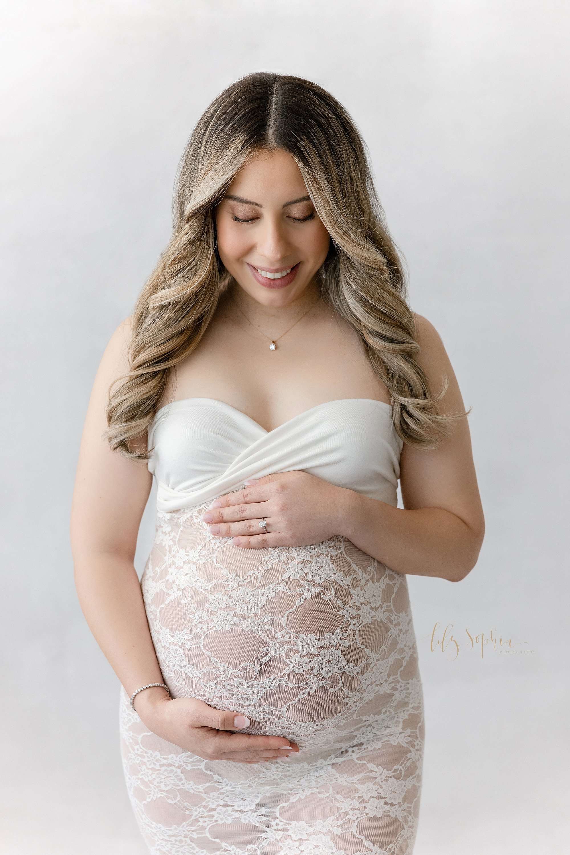 Maternity portrait of an expectant mother wearing a lace gown with a jersey knit strapless bodice as she looks down at her womb while framing it with her hands taken using natural light in a photography studio near Ansley Park in Atlanta. 