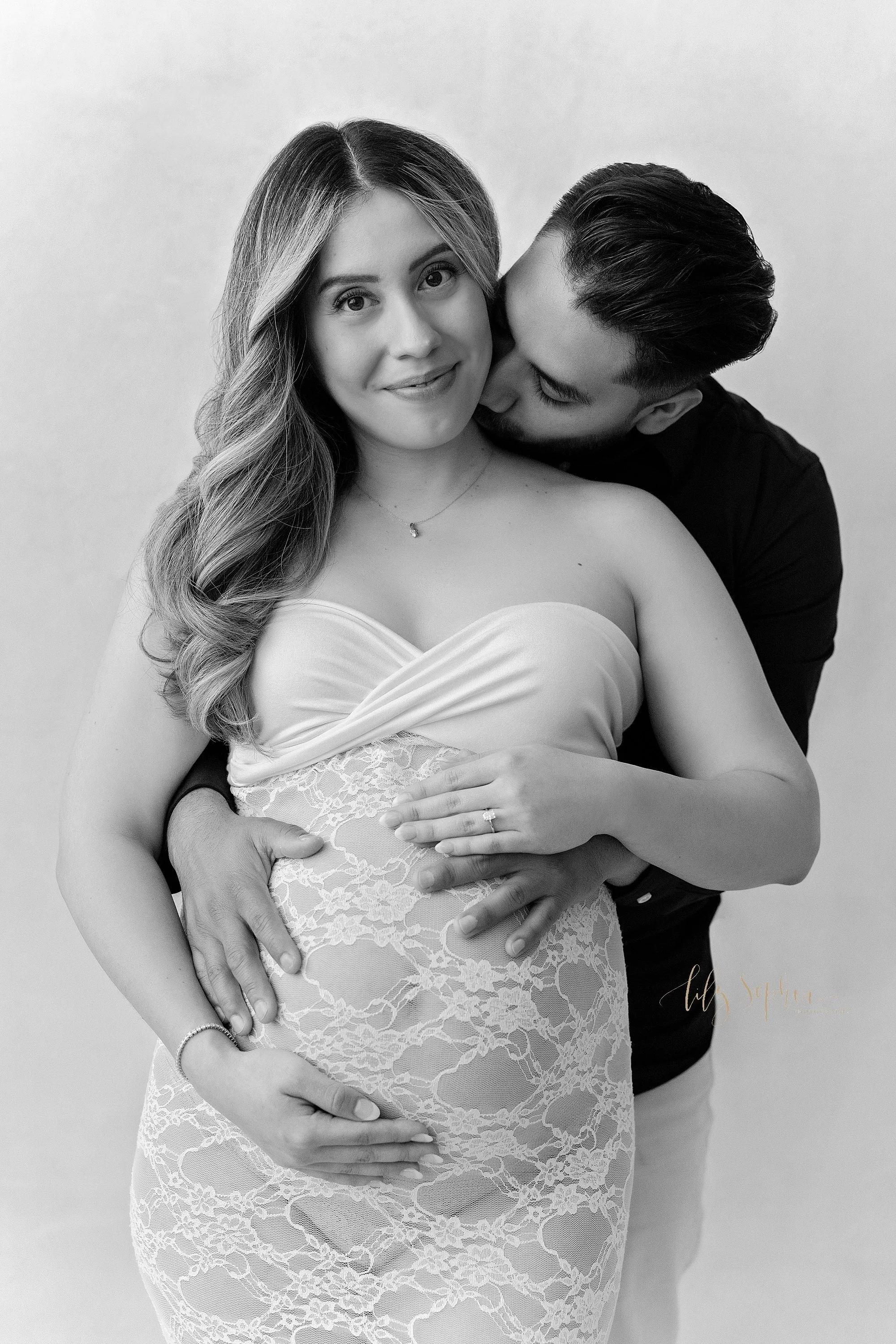  Black and white maternity photo of a pregnant mother wearing a strapless knit bodice lace gown as her husband stands behind her to reach his hands to place them on their child in utero while the mother holds the base of her womb with her right hand 