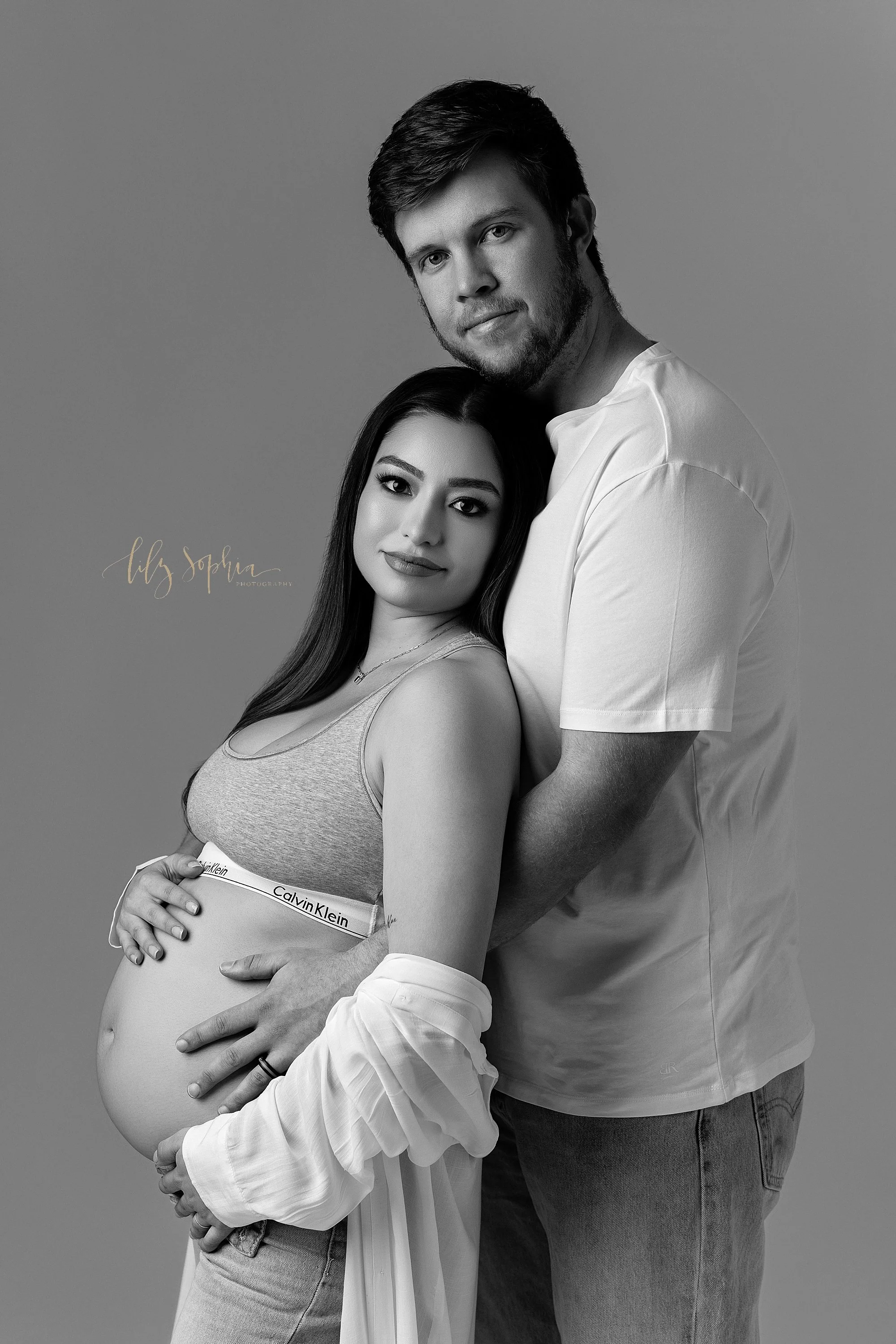  An expectant mother stands with her back against her husband’s chest as he stands behind her and places his hands on his child in utero as his wife holds the base of her bare womb with her right hand while the couple looks over their right shoulders