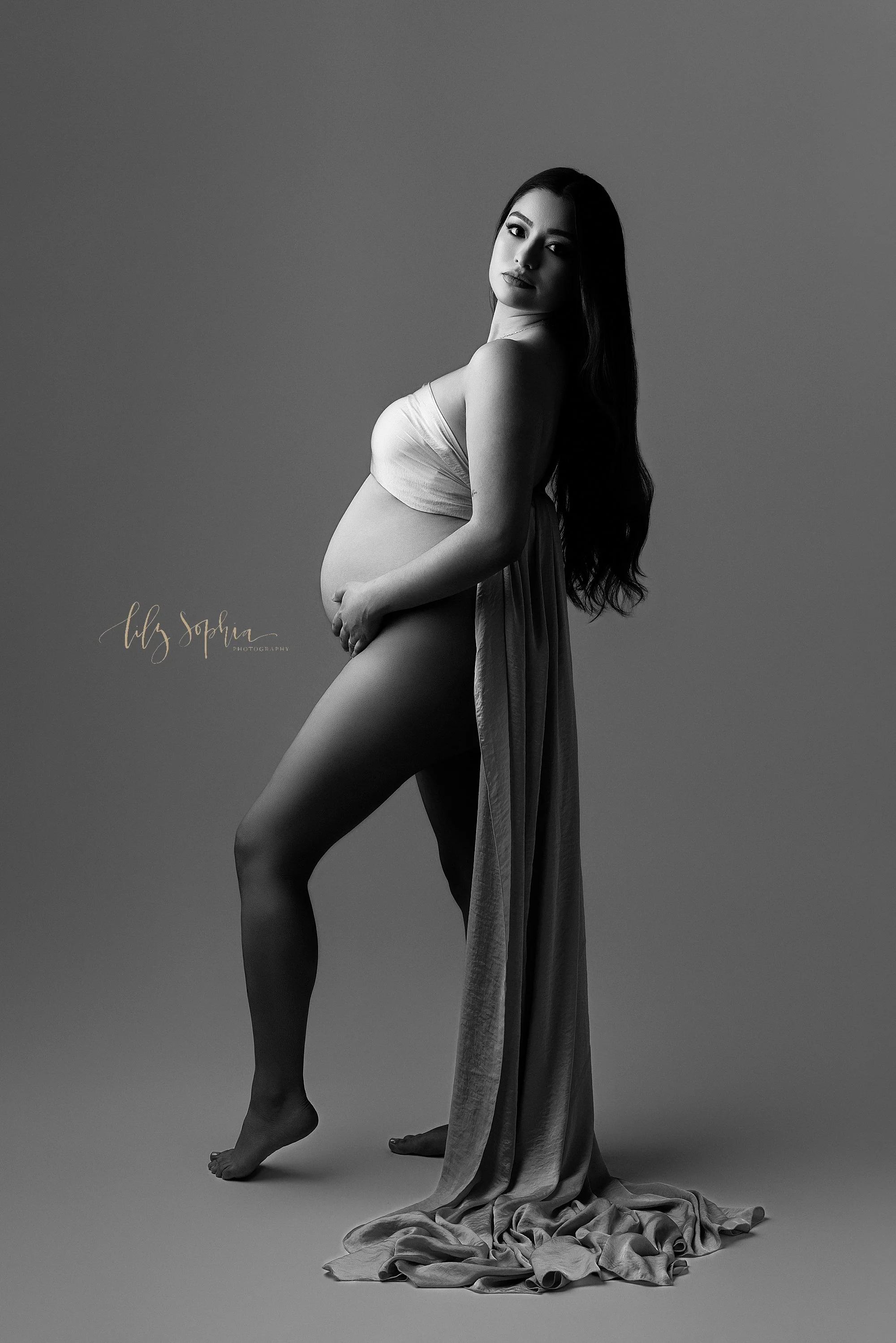  Dramatic black and white portrait of an expectant mother with her breast draped with a silky piece of fabric as she bends her right leg to stand on tiptoe and holds her bare womb while looking over her right shoulder taken near Midtown in Atlanta du