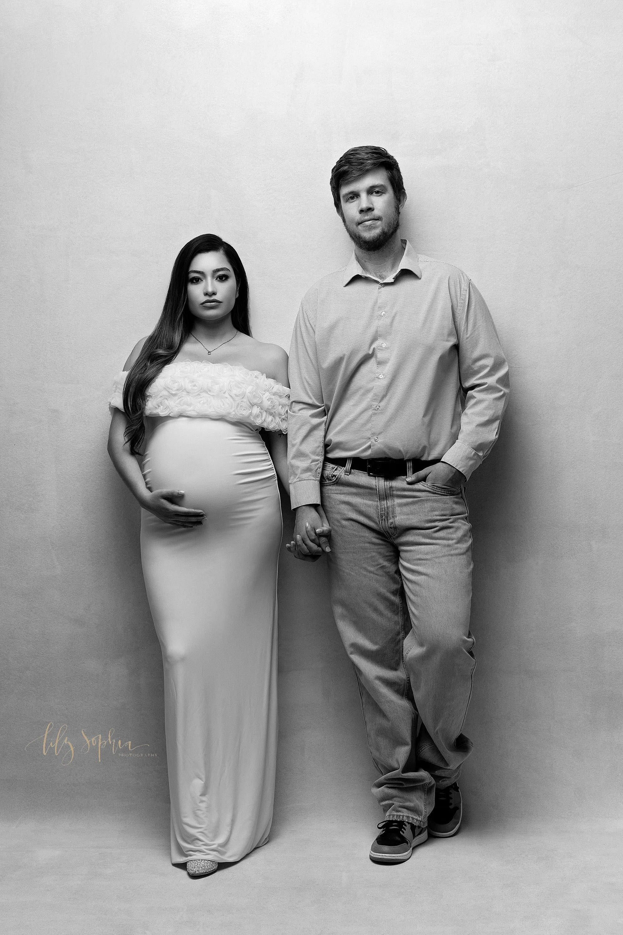  Black and white modern maternity photo of an expectant couple standing with their backs to a wall and holding hands as the mother holds the base of her womb with her right hand taken near Decatur in Atlanta, Georgia. 