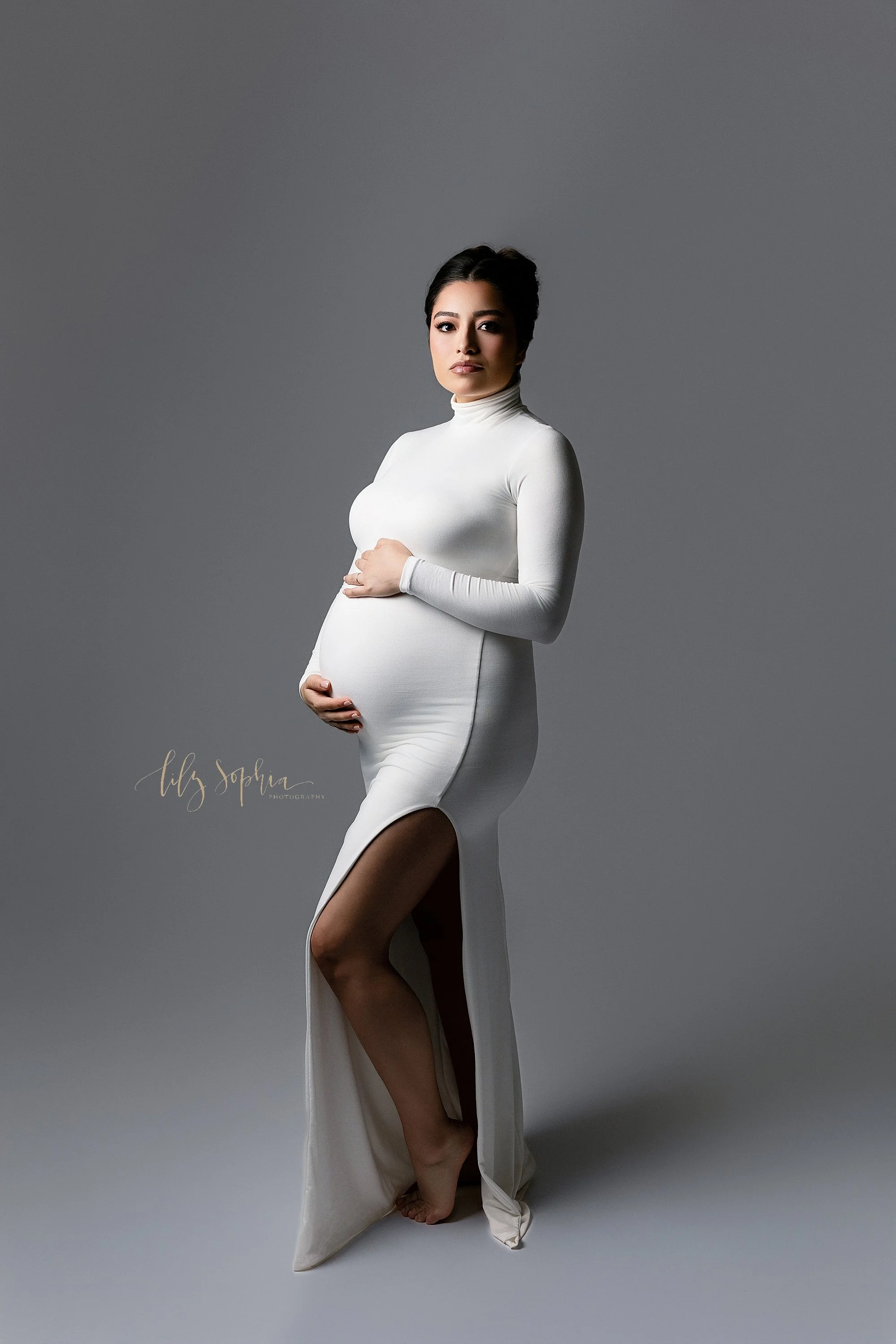  Modern maternity photography session of an expectant mother as she stands wearing a white side split long sleeved turtleneck knit gown as she frames her womb with her hands and looks toward her right shoulder and bends her right leg taken near Morni