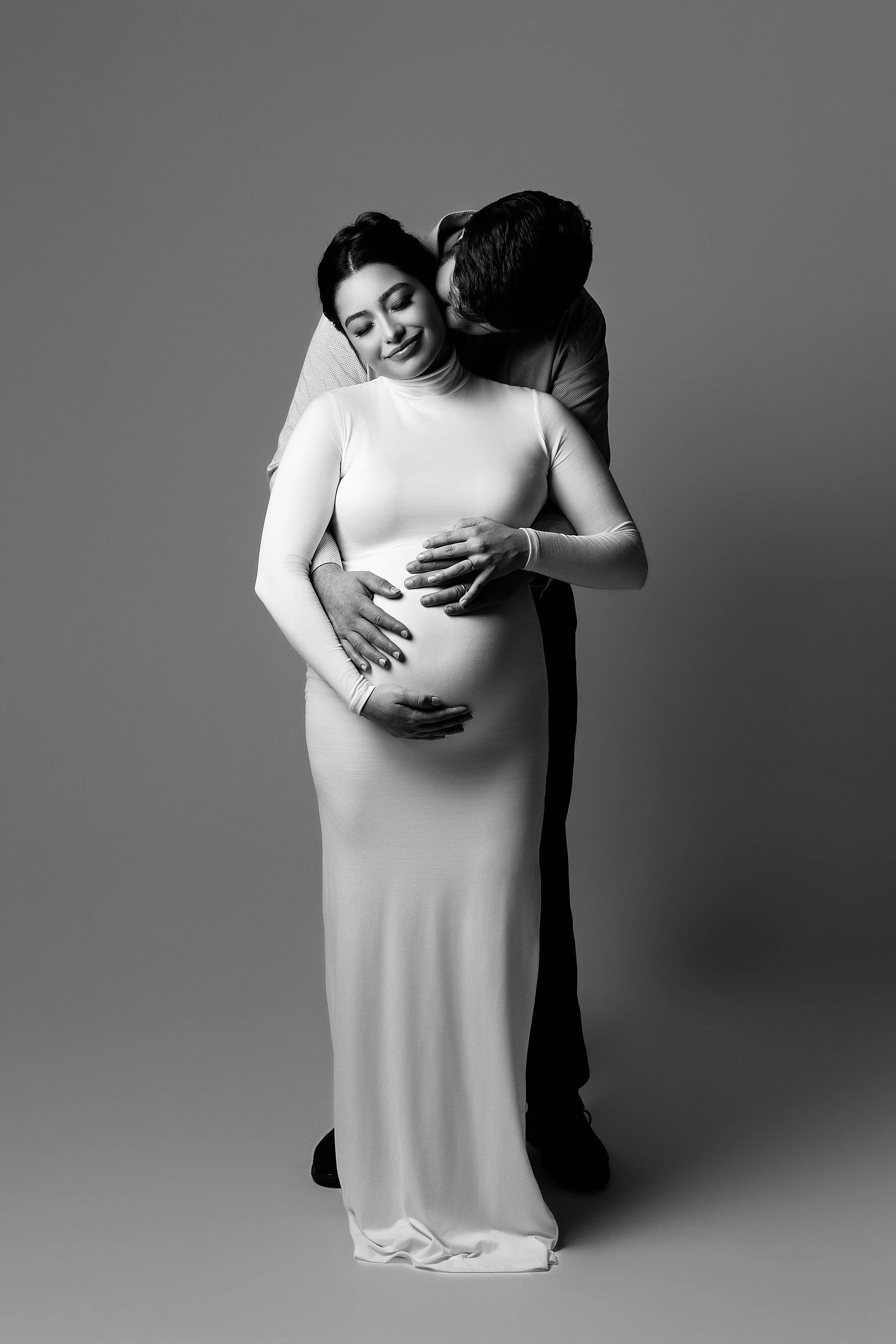  The love of a mother and father is featured during this modern maternity photo session as the expectant mother stands in front of her husband with her back to his chest as he reaches his hands to hold his child in utero and kisses the neck of his wi