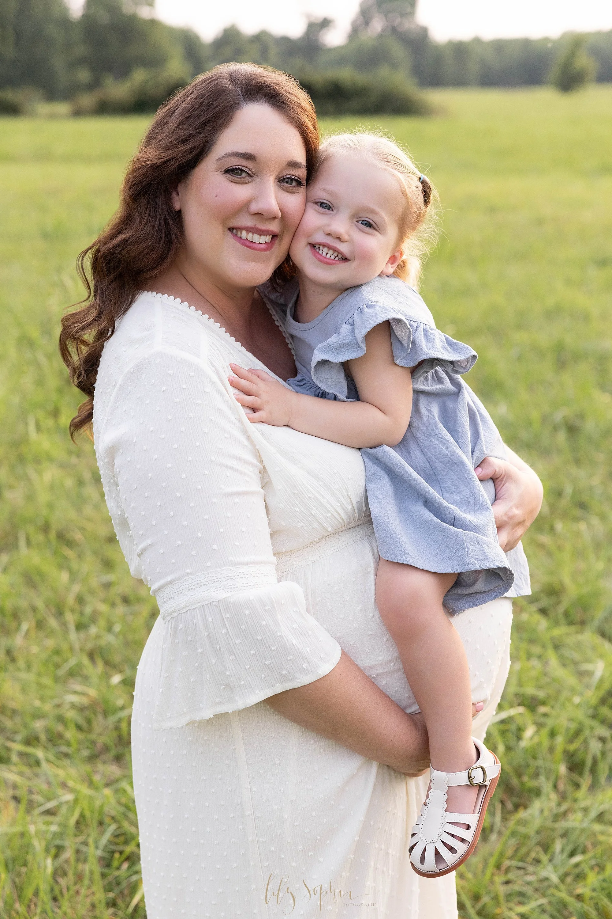  First year collection for a maternity session taken at sunset in an Atlanta field of a pregnant mother holding her young daughter as mom holds the base of her womb with her right hand and hugs her daughter to her chest with her right hand. 