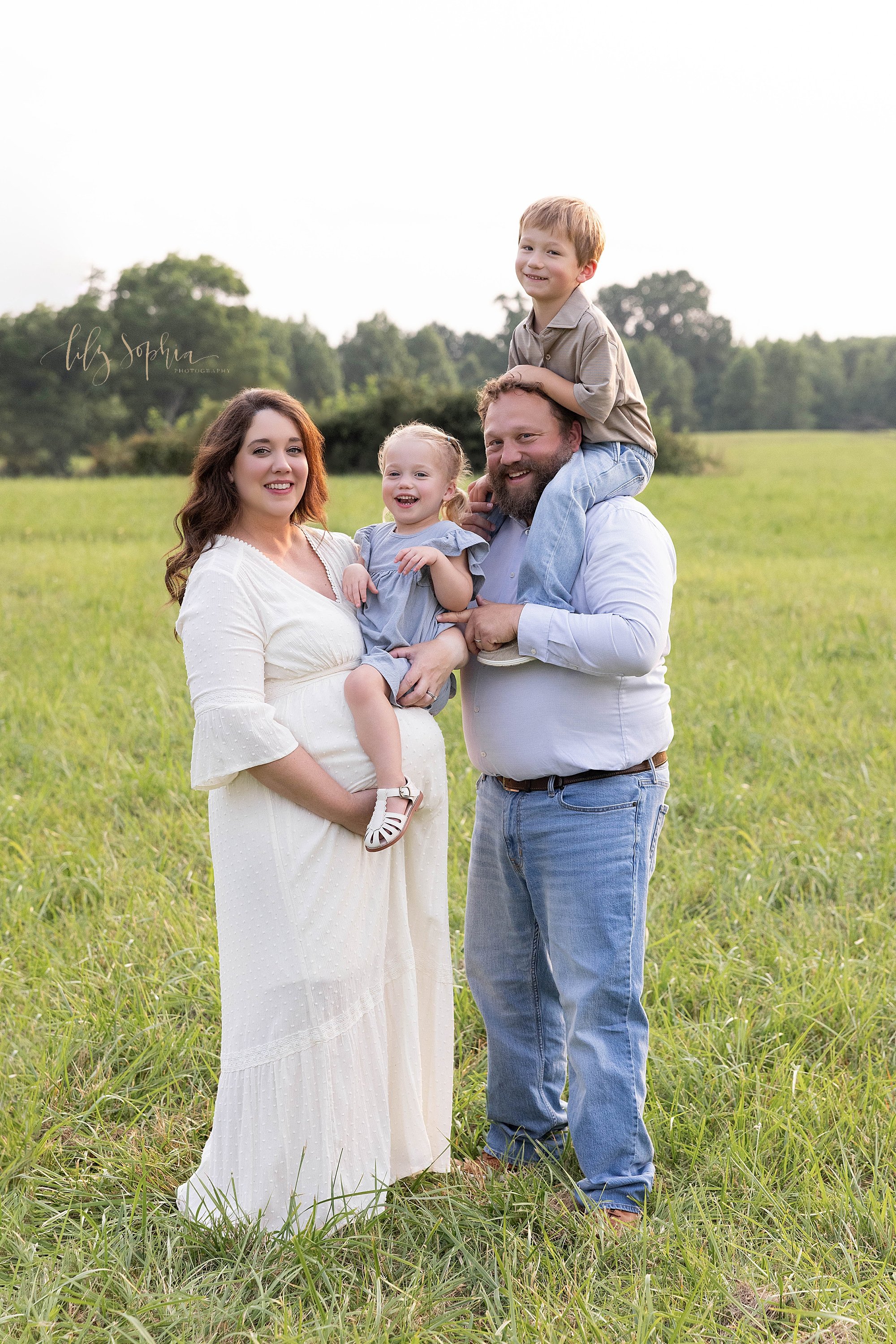 Family maternity first year collection session with a father holding his young son on his shoulders as his pregnant wife stands facing him as she holds their young daughter while the family stands in a field near Atlanta, Georgia at sunset. 