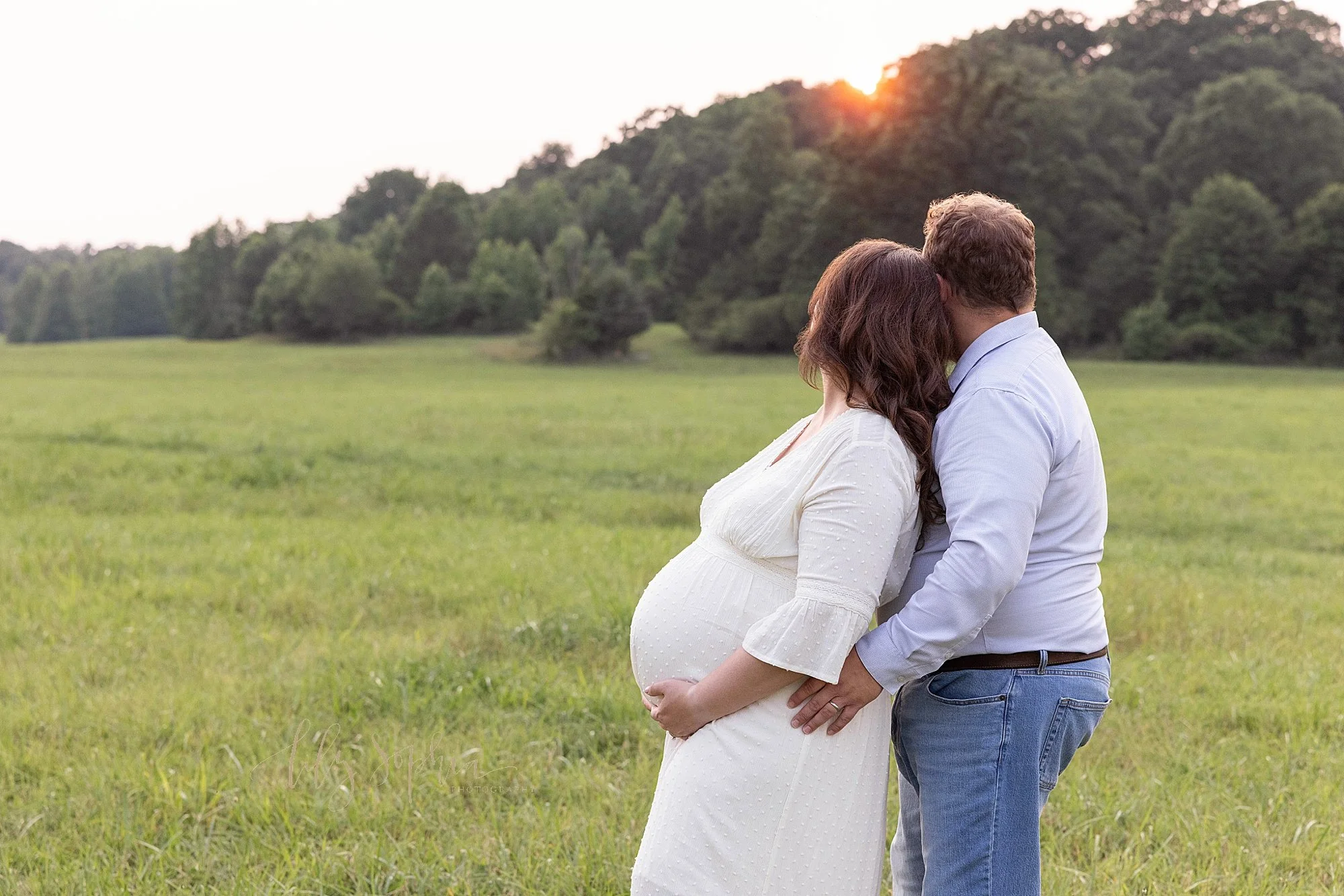  Profile maternity portrait of a mother holding the base of her womb as she stands with her back to her husband’s chest as the couple looks over their right shoulders to look to the future as the sunset is seen in the field near Atlanta, Georgia. 
