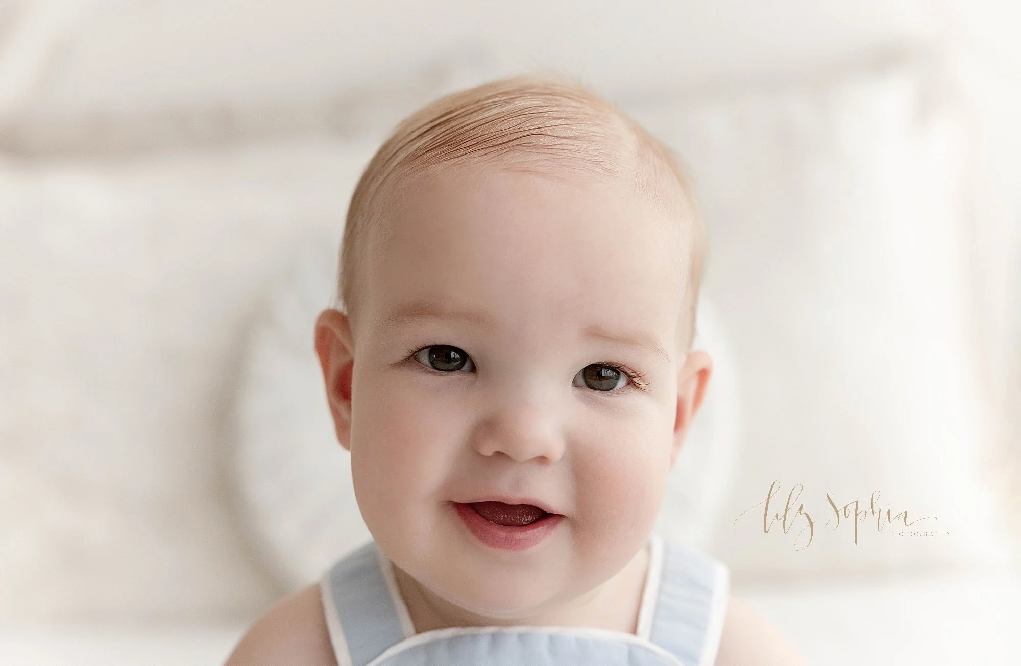  Close-up portrait of a one year old baby boy as he sits on a bed for his first birthday photography session in a natural light studio near Kirkwood in Atlanta, Georgia. 