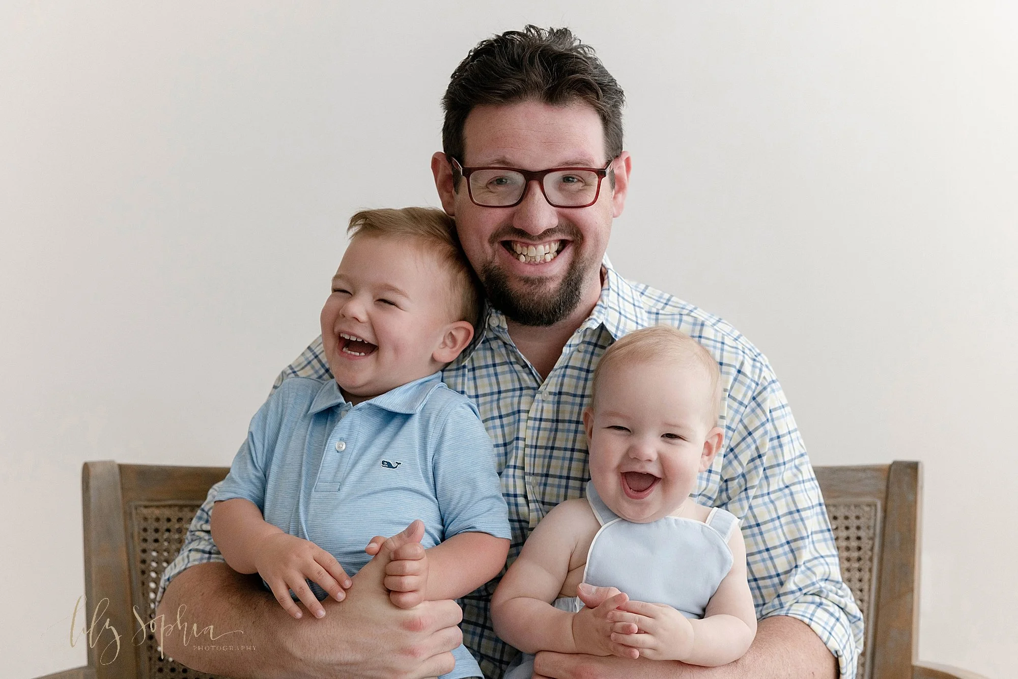  A fun family photo of a dad as he sits on a cane backed bench with his sons on his lap as he tickles them and they laugh taken near Ansley Park in Atlanta in a photography studio that uses natural light. 